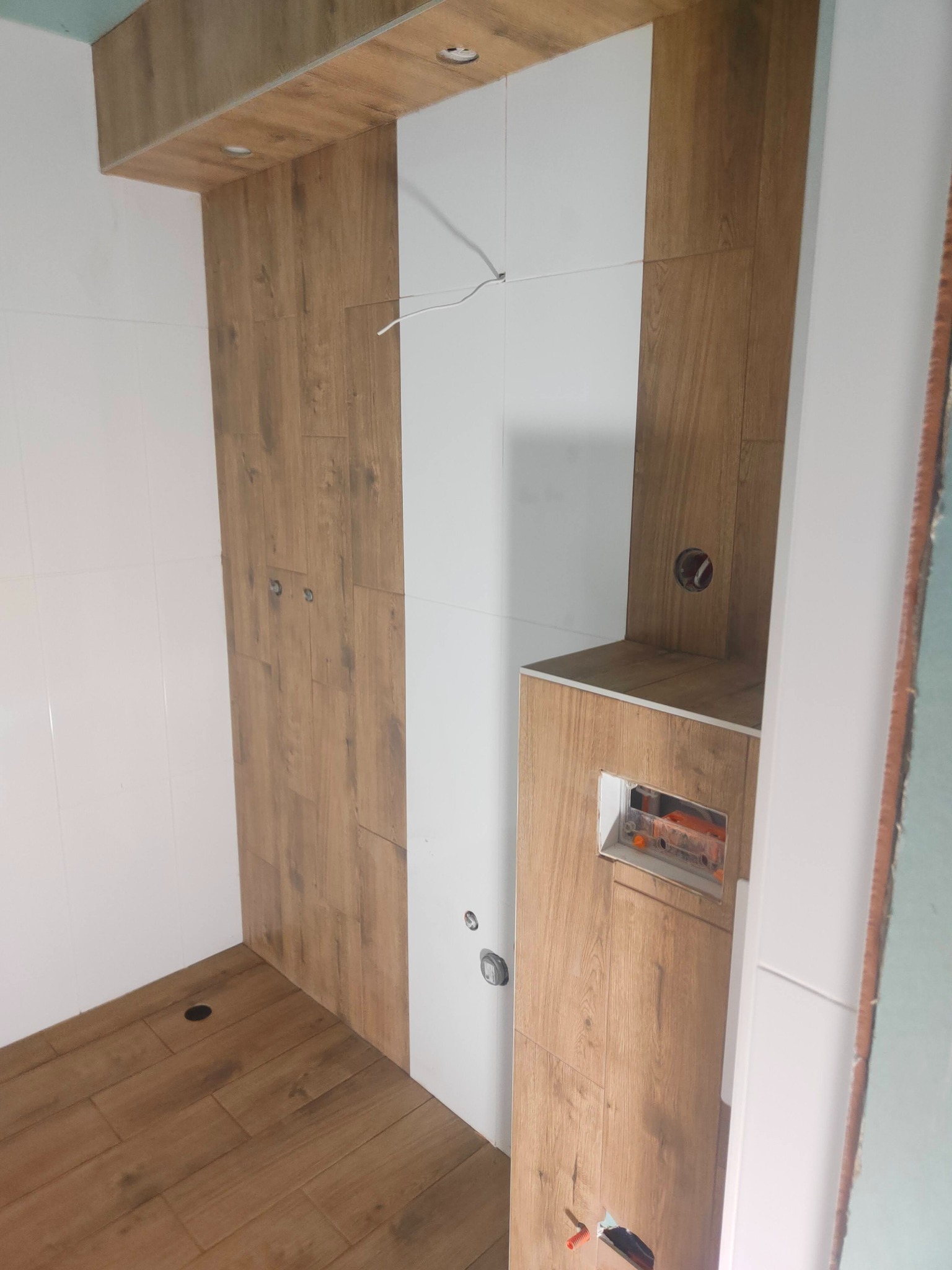 Wnętrze łazienki w trakcie prac wykończeniowych. Widoczne płytki imitujące drewno na ścianach i podłodze, białe płytki oraz wystające przewody elektryczne.