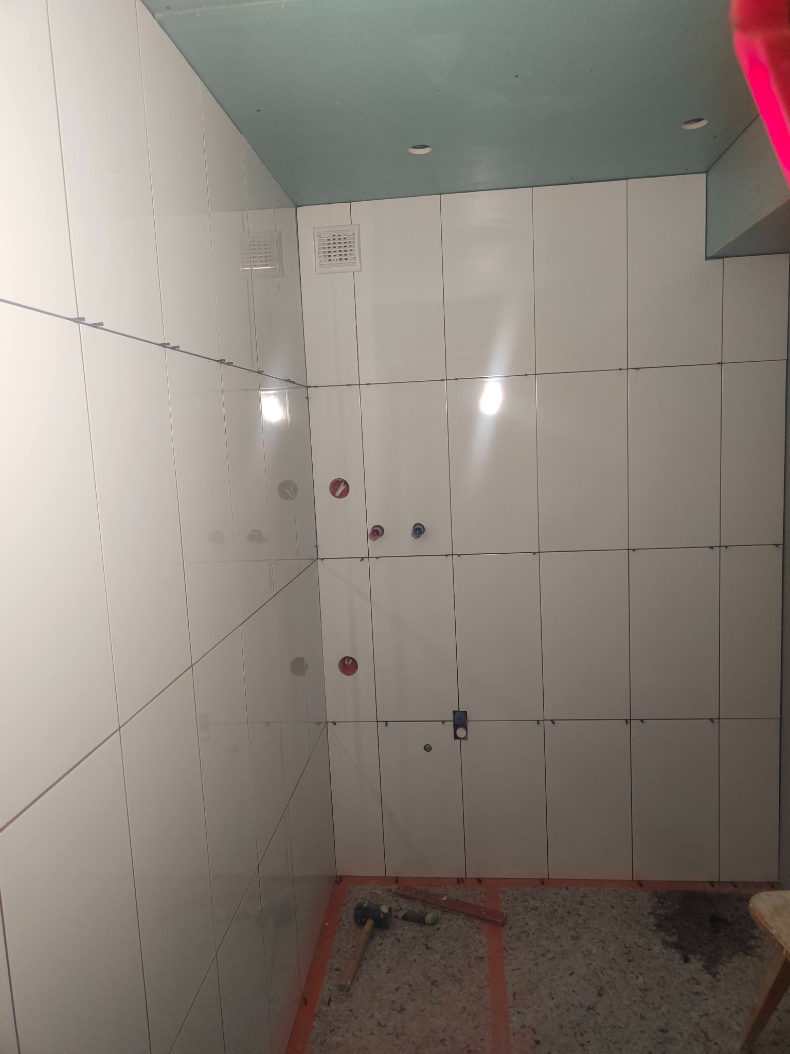 Białe płytki ścienne w trakcie układania w rogu łazienki. Widoczne krzyżyki dystansowe, otwory na instalacje i narzędzia na podłodze. Sufit pomalowany na zielono.