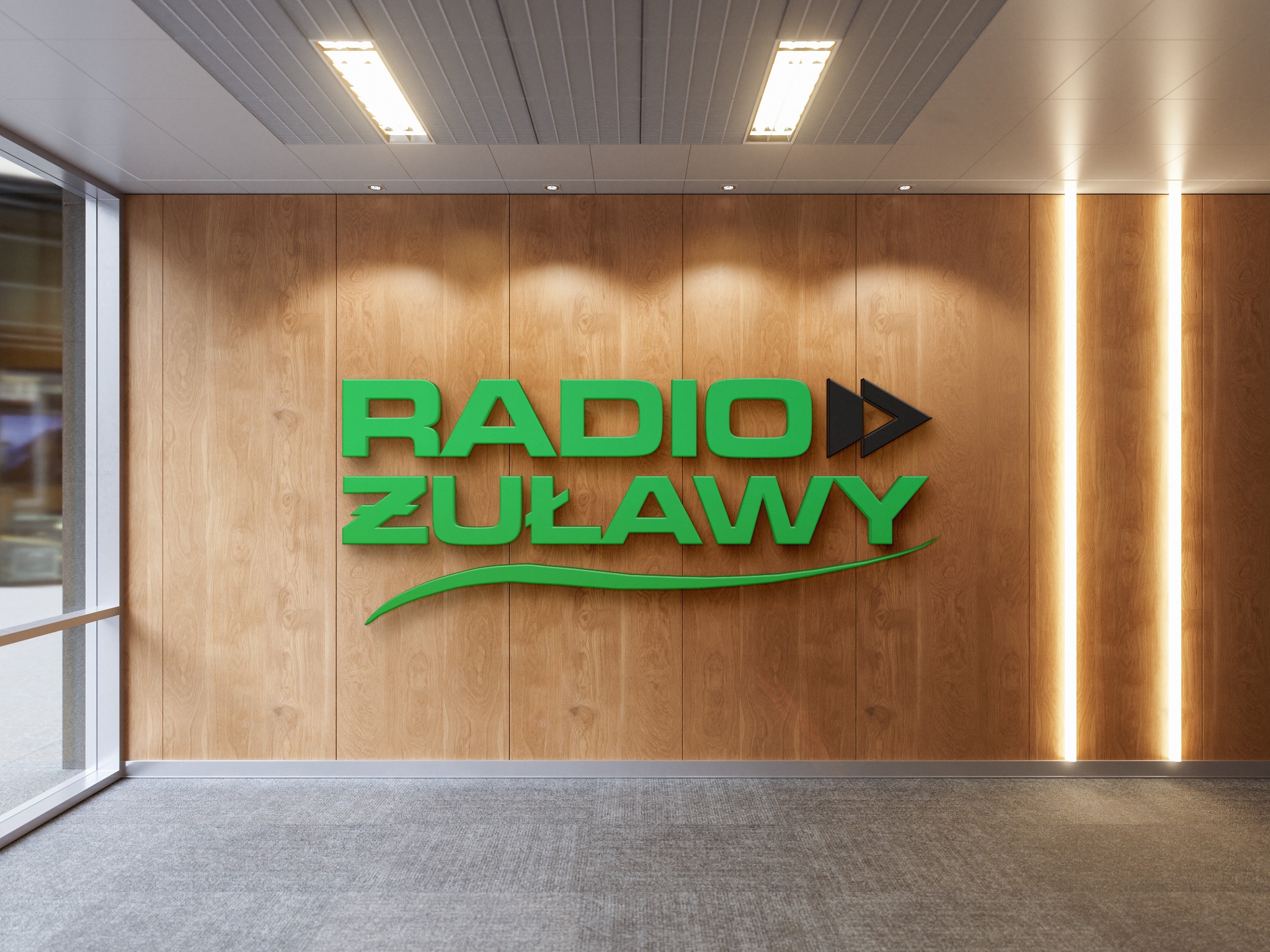 Zielone logo 'RADIO ŻUŁAWY' z czarną strzałką, umieszczone na drewnianej ścianie w nowoczesnym biurze z szarym dywanem i oświetleniem LED.