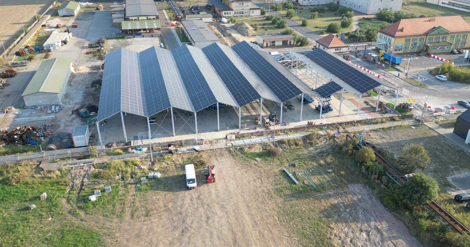 Widok z lotu ptaka na dużą konstrukcję z panelami słonecznymi, w tle budynki przemysłowe i teren budowy z zaparkowanymi pojazdami.