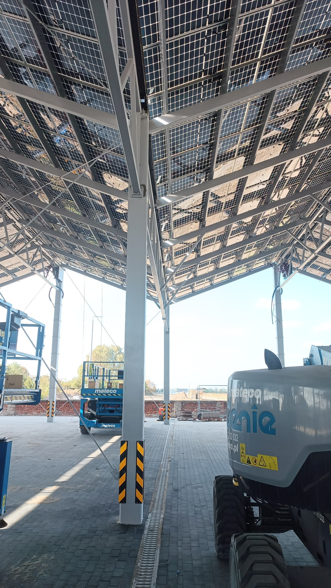 Montaż paneli fotowoltaicznych na konstrukcji stalowej z użyciem podnośników koszowych, widok z dołu na panele i konstrukcję.