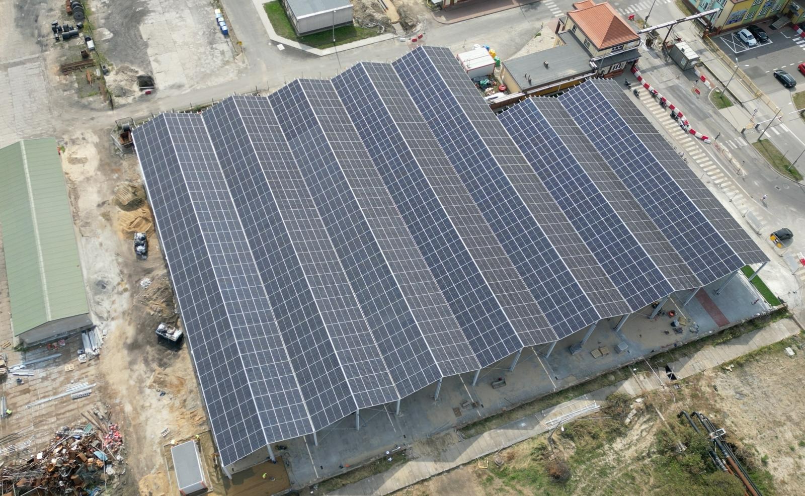 Widok z góry na dużą konstrukcję z panelami słonecznymi o falującym układzie, wspartą na metalowych kolumnach, otoczoną placem budowy z piaskiem i materiałami, w tle zabudowania miejskie i ulica.