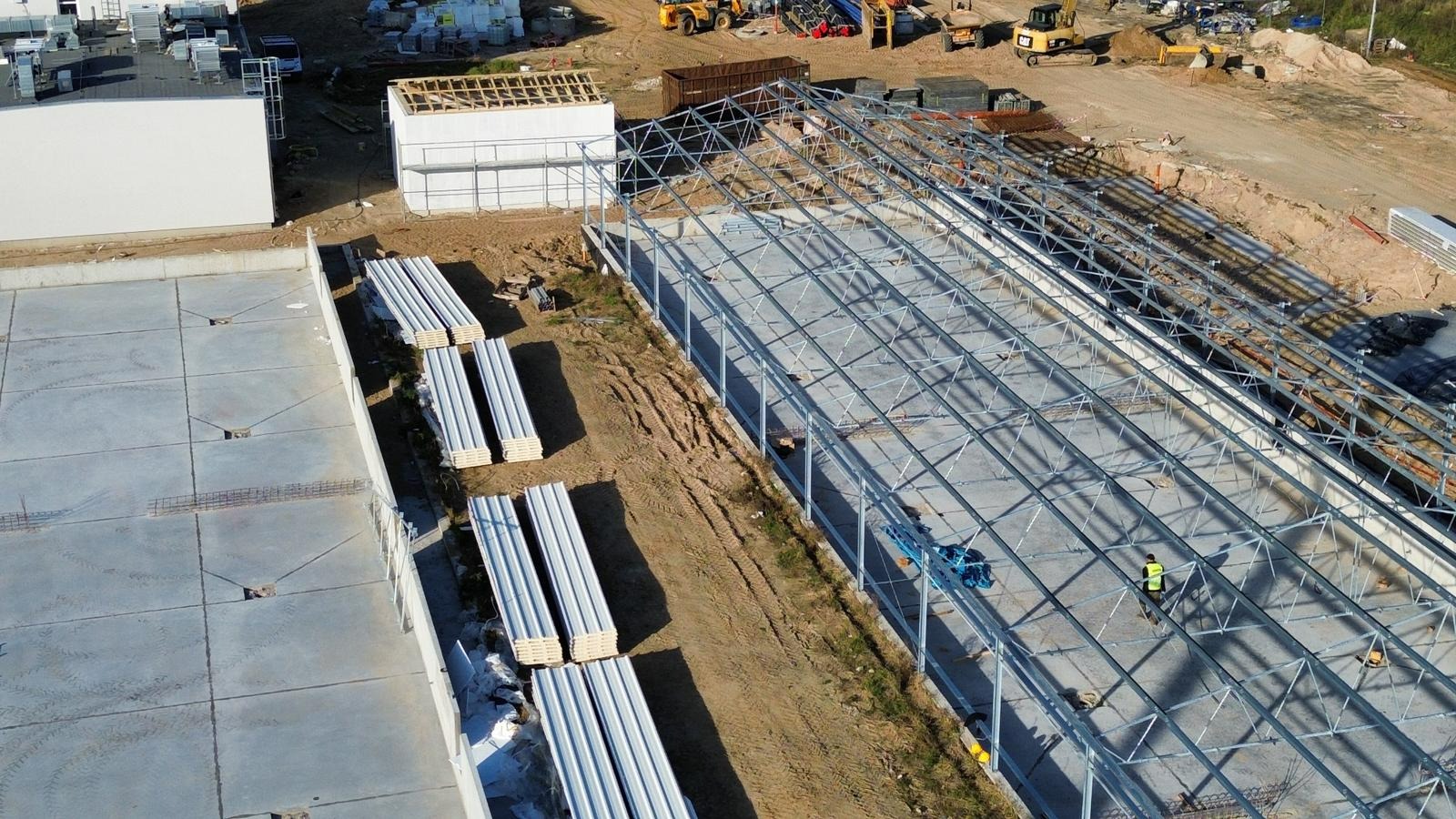 Widok z góry na plac budowy z konstrukcją stalową hali w trakcie montażu, widoczne przygotowane płyty warstwowe, ciężki sprzęt budowlany i pracownik w kamizelce odblaskowej.