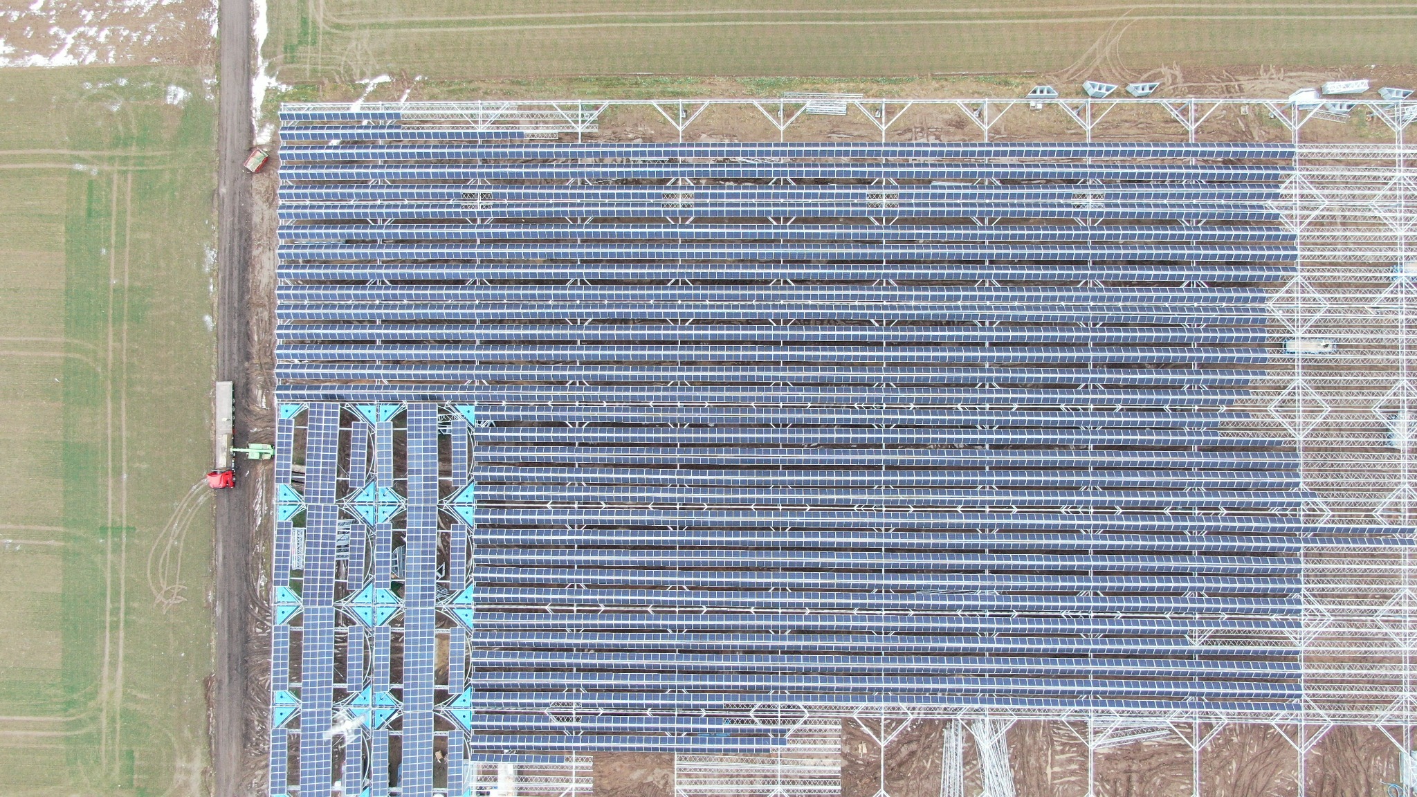 Widok z lotu ptaka na rozległą farmę paneli słonecznych w trakcie instalacji, z widocznym traktorem i przyczepą na skraju pola, częściowo pokrytego śniegiem.