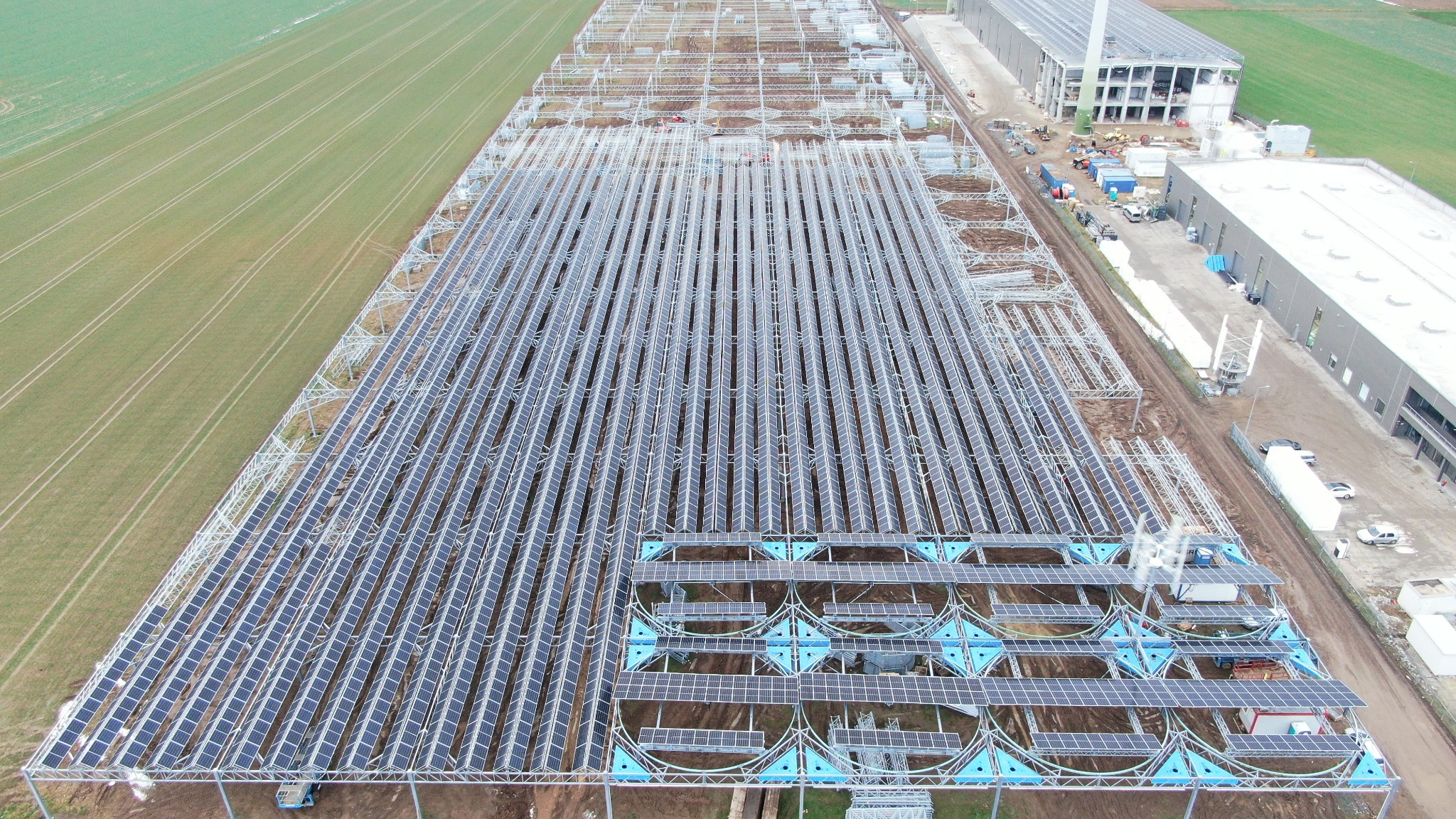 Widok z lotu ptaka na rozległą farmę paneli słonecznych w trakcie budowy, z widocznymi konstrukcjami wsporczymi i budynkami przemysłowymi w tle.