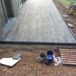 Nowoczesny taras z szarej kostki brukowej ułożony przy budynku, widoczne narzędzia i materiały budowlane w trakcie prac wykończeniowych.