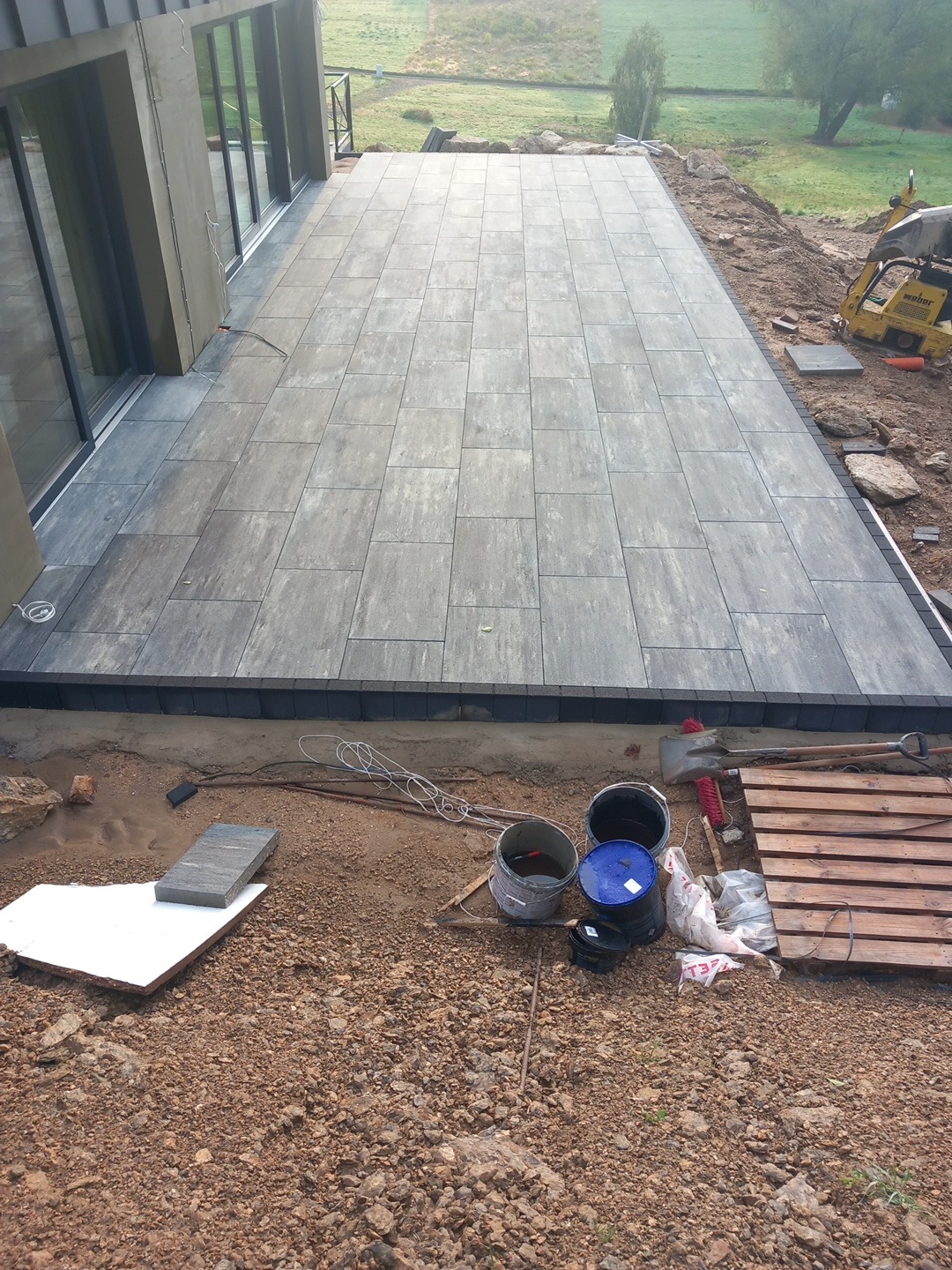 Nowoczesny taras z szarej kostki brukowej ułożony przy budynku, widoczne narzędzia i materiały budowlane w trakcie prac wykończeniowych.