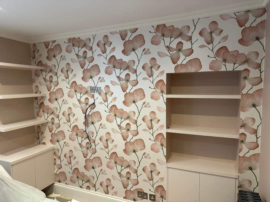 Pokój z kwiatową tapetą w odcieniach różu i beżu, zabudowanymi półkami w kolorze ścian oraz niedokończonymi detalami wykończeniowymi przy listwach podłogowych.