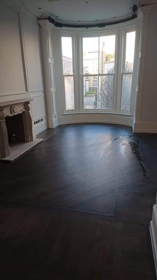 Pokój z ciemną, świeżo lakierowaną parkietową podłogą ułożoną w jodełkę, widoczne ślady lakieru, biały kominek i duże okno wykuszowe z widokiem na ulicę.