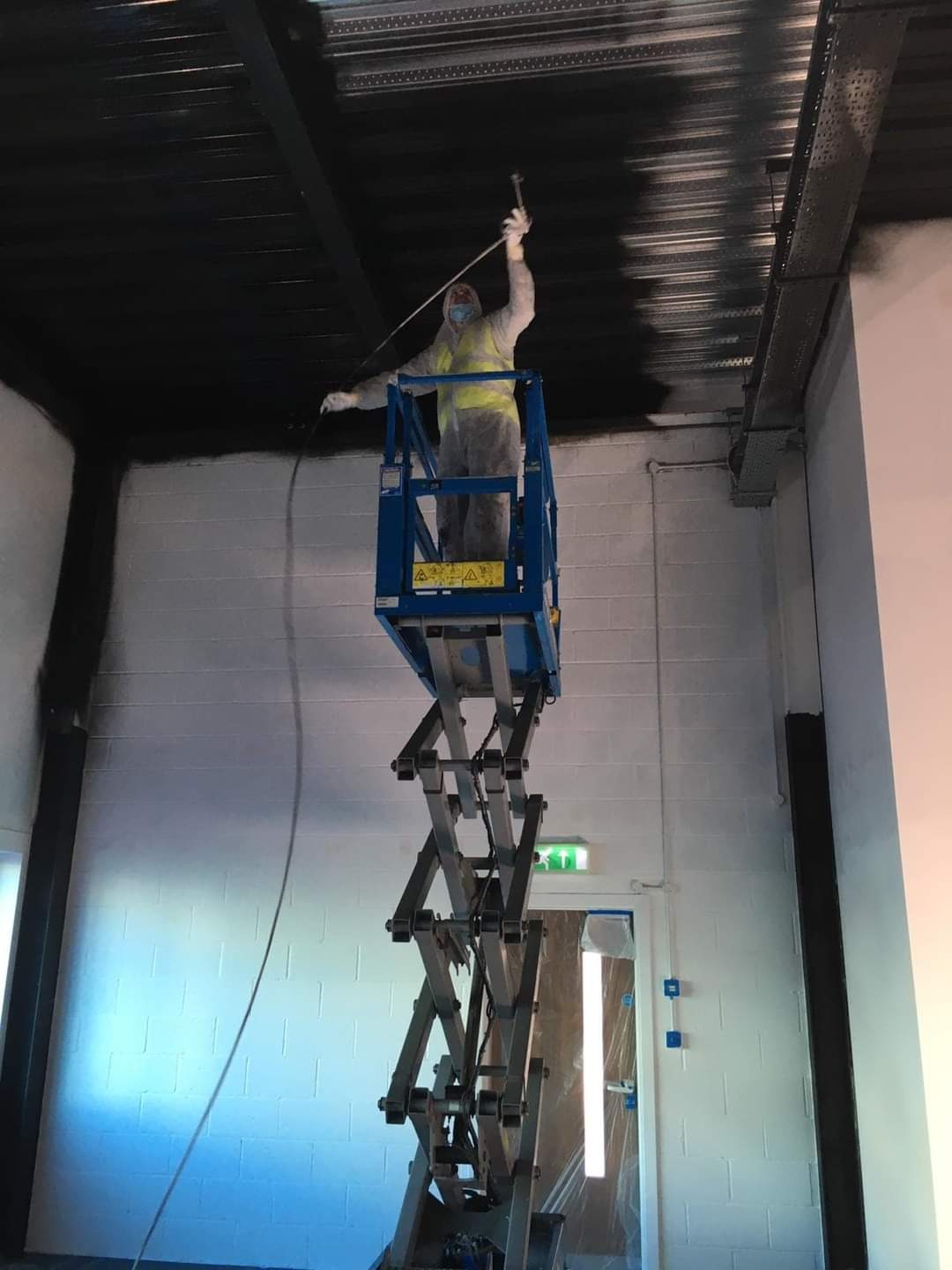 Pracownik w kombinezonie ochronnym na podnośniku nożycowym maluje sufit natryskowo na czarno, widoczne świeżo pomalowane fragmenty i instalacje pod sufitem.