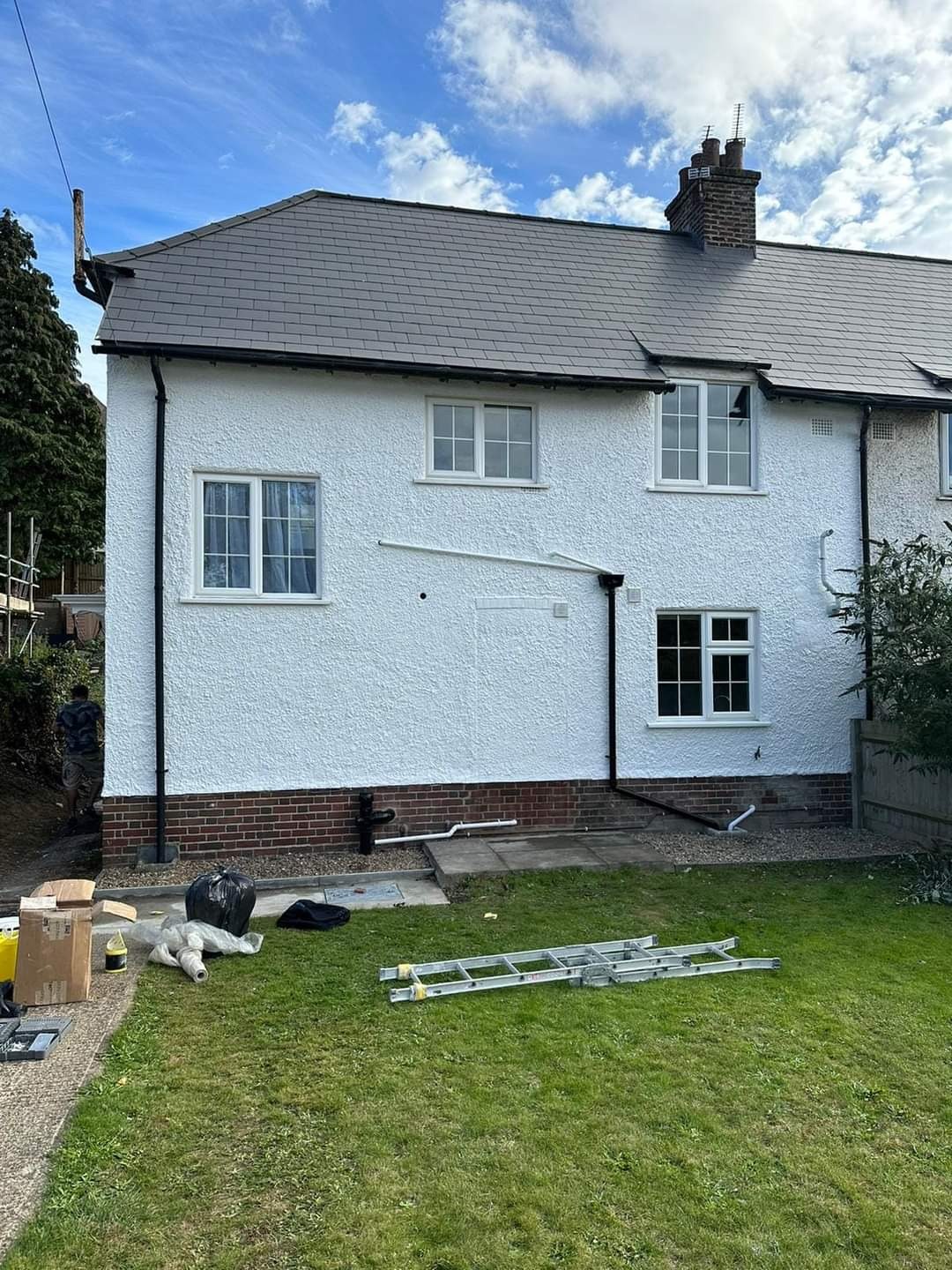Odnowiona elewacja domu z białym tynkiem strukturalnym, nową szarą dachówką i białymi oknami, widoczne narzędzia i materiały budowlane na trawniku.