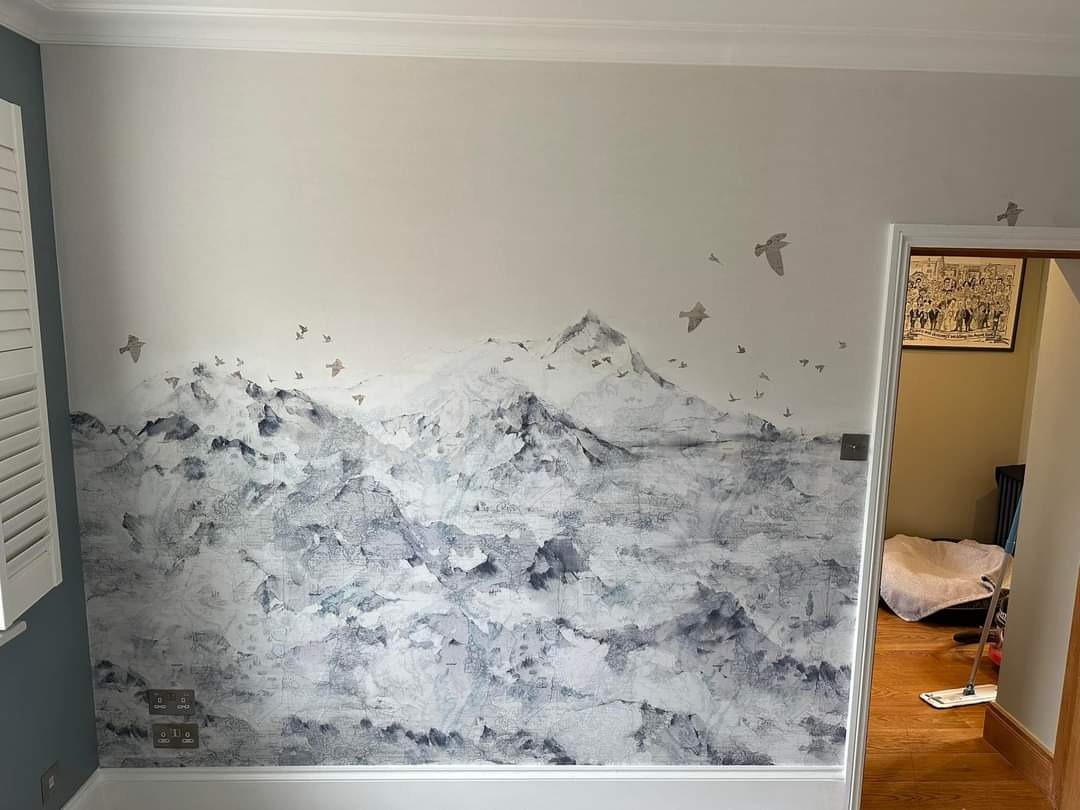 Ściana z ręcznie malowanym motywem górskim i ptakami w szaro-białej tonacji, widoczne gniazdka elektryczne i fragment parkietu.