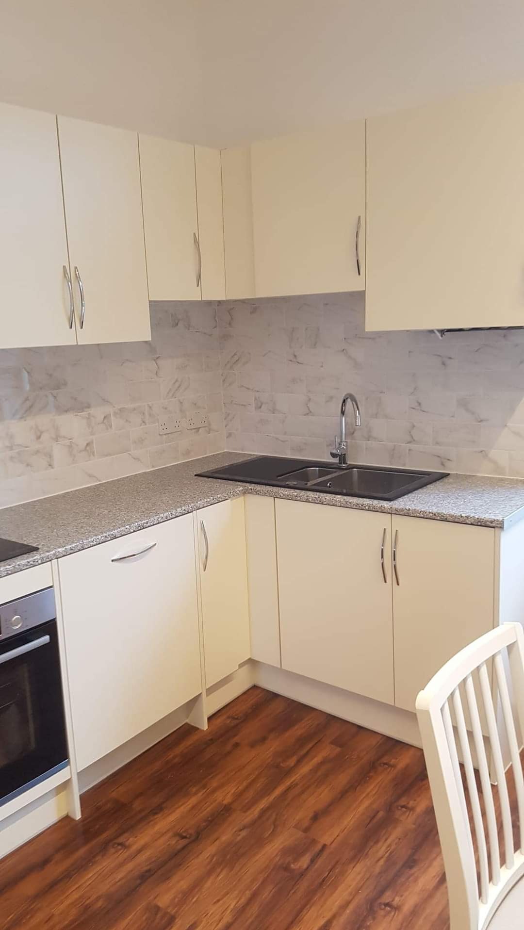 Jasna zabudowa kuchenna z marmurowym wzorem na ścianie, ciemny zlew i drewniana podłoga w odcieniach brązu.