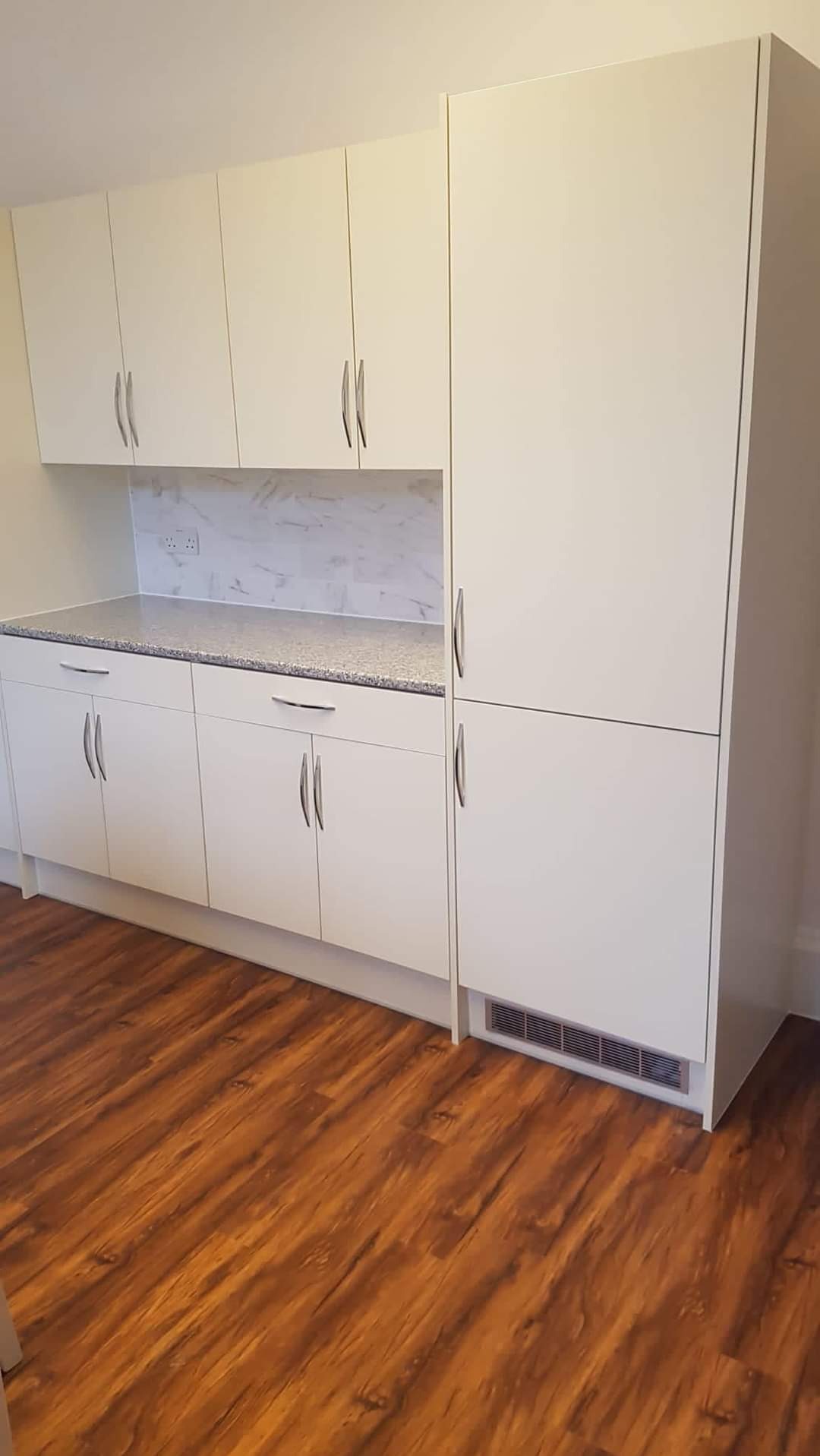 Biała zabudowa kuchenna z szafkami górnymi i dolnymi, blatem w kolorze soli z pieprzem oraz brązową podłogą imitującą drewno.