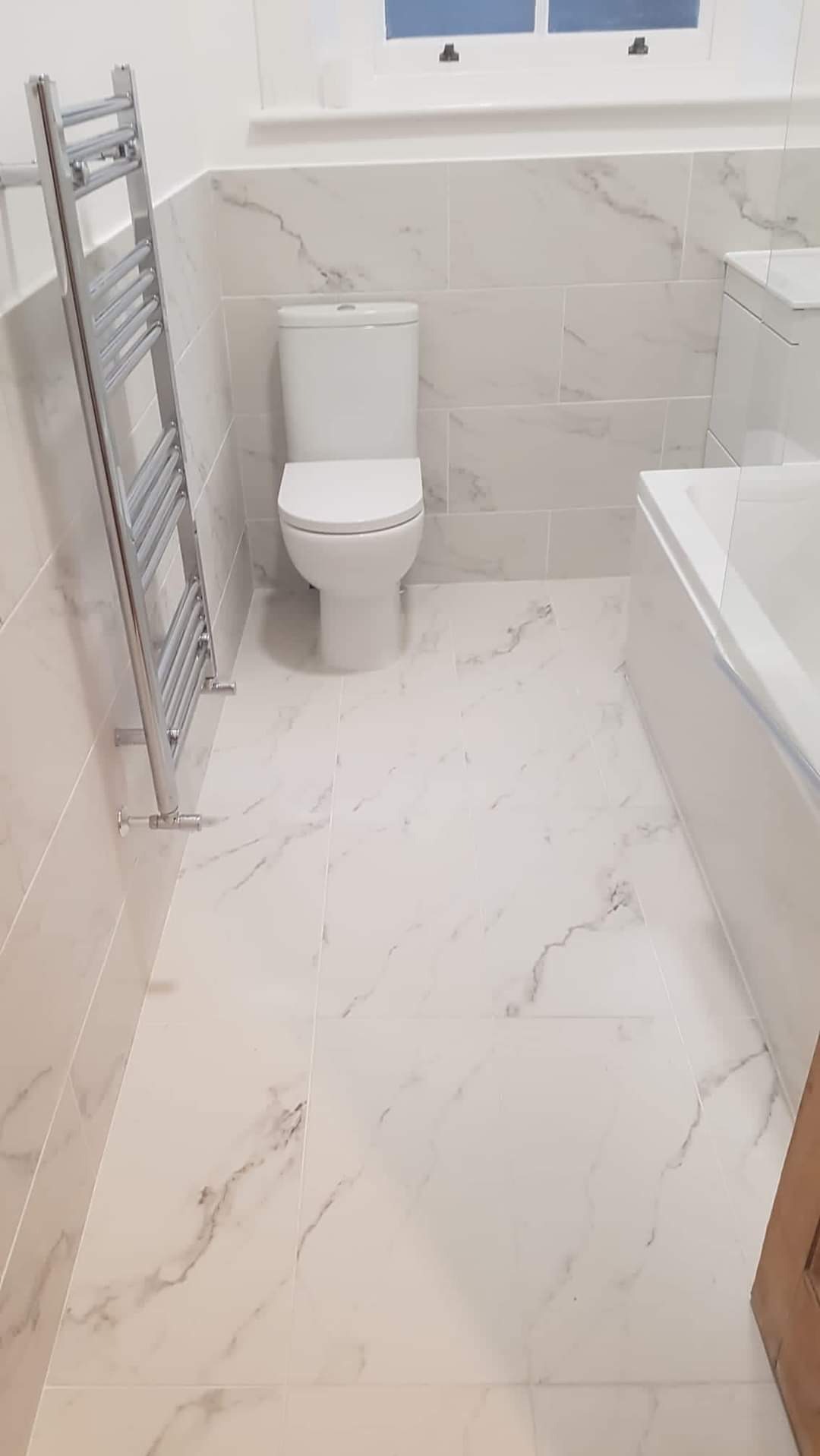 Jasna łazienka z marmurowymi płytkami, widoczny grzejnik drabinkowy, toaleta i fragment wanny, oświetlona przez okno.