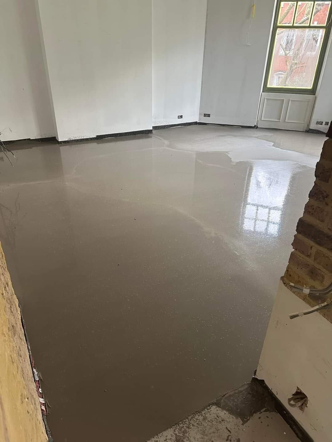 Wylewka samopoziomująca na podłodze w jasnym pomieszczeniu, widoczne ślady po narzędziach i odbicie okna w mokrej powierzchni, fragment ceglanej ściany z instalacją elektryczną w prawym dolnym rogu.