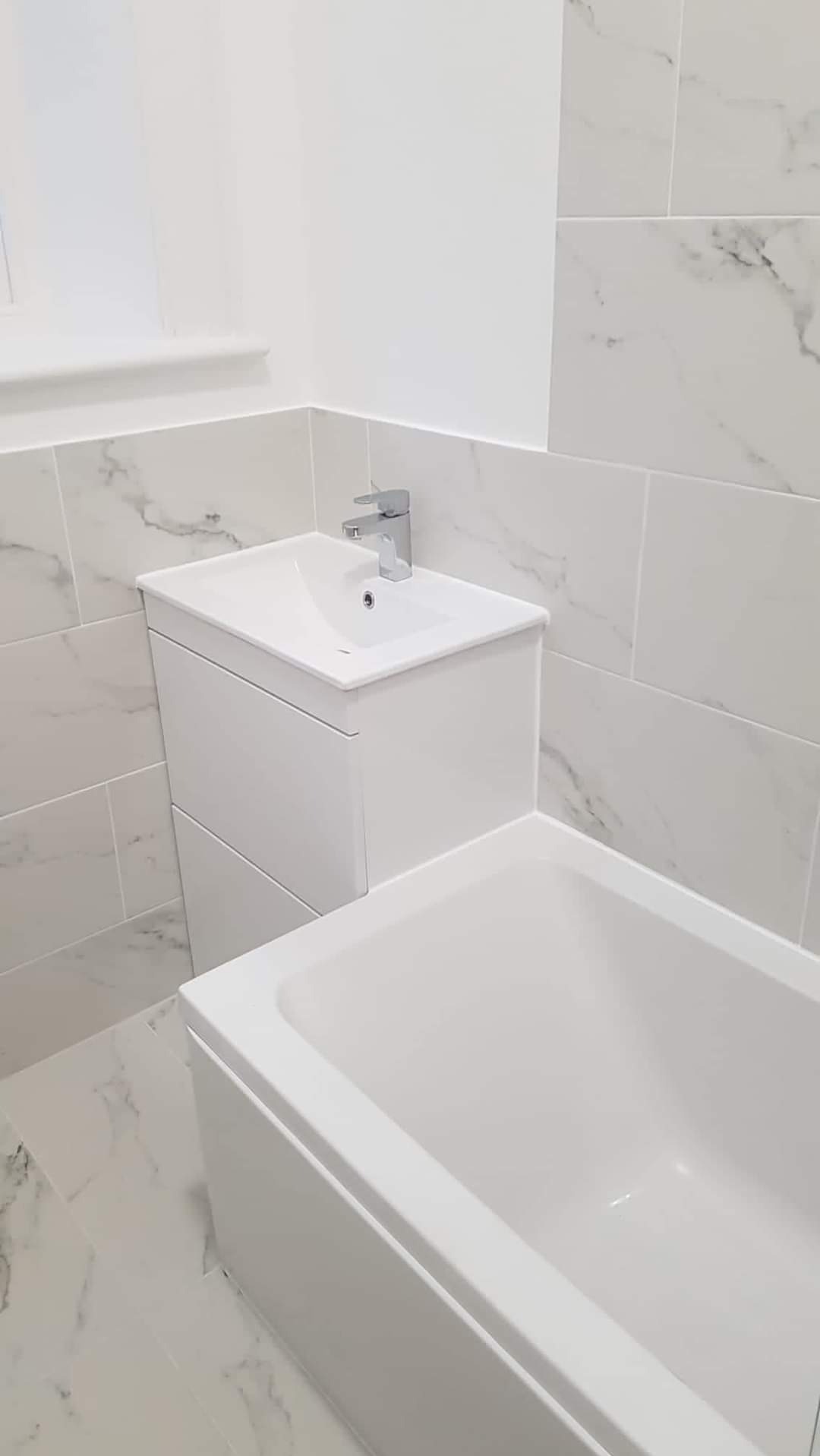 Nowoczesna łazienka w bieli: umywalka z szafką, bateria chromowana i wanna, wykończona płytkami imitującymi marmur z delikatnymi szarymi żyłkami.