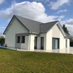 Nowoczesny, parterowy dom z szarą dachówką i jasną elewacją, otoczony trawnikiem i częściowo wyłożony płytami.