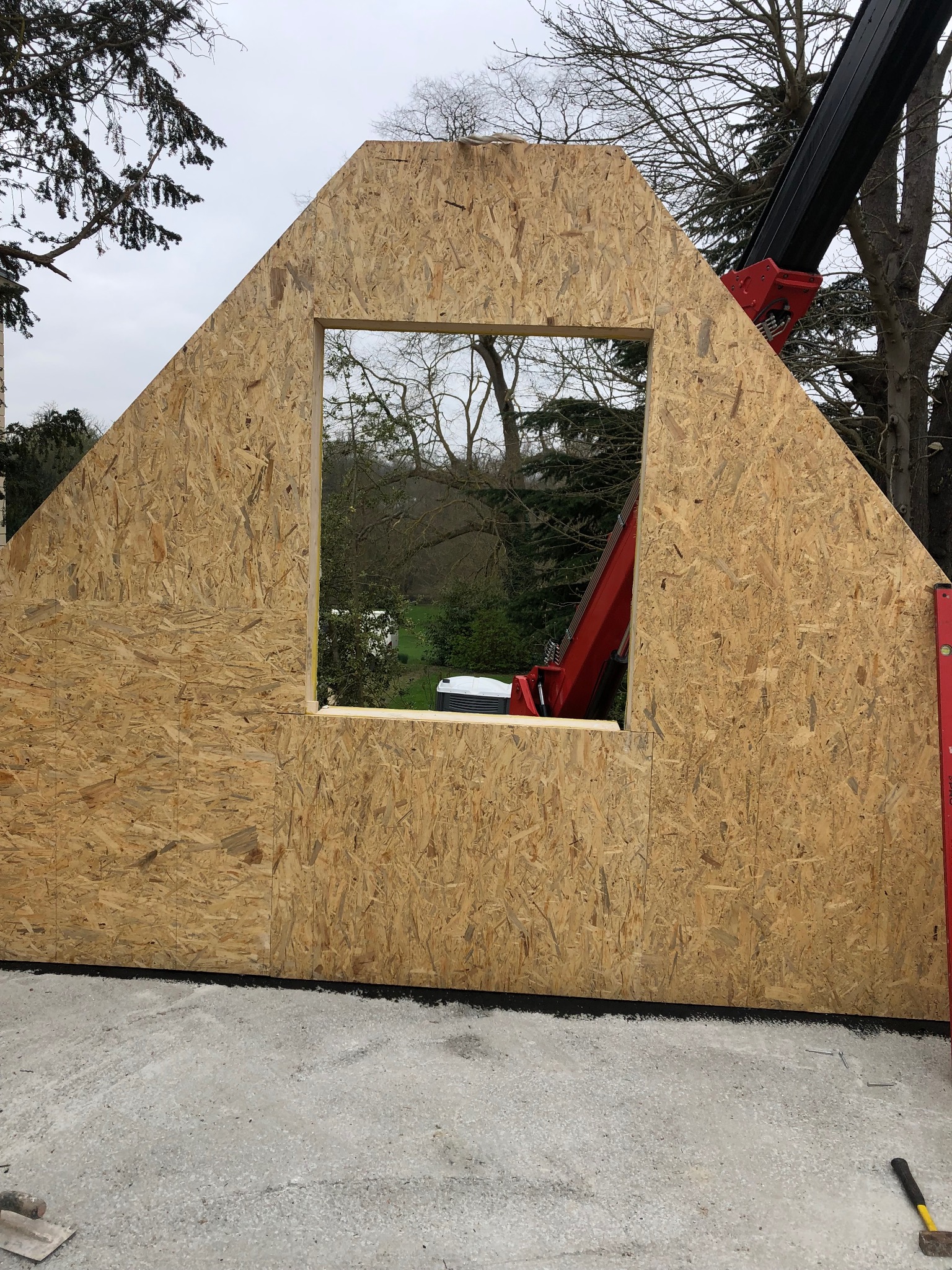 Montaż ściany z płyty OSB z otworem okiennym, podnoszonej przez czerwony dźwig na tle zieleni.