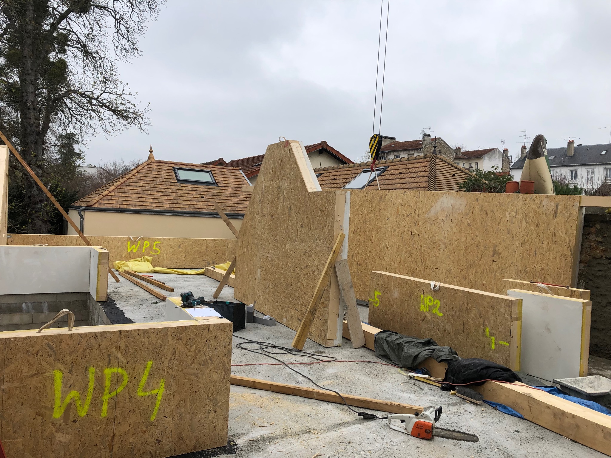 Montaż ścian z płyt OSB z oznaczeniami WP4 i WP5 na fundamentach, widoczny dźwig i narzędzia budowlane, w tle zabudowa jednorodzinna.