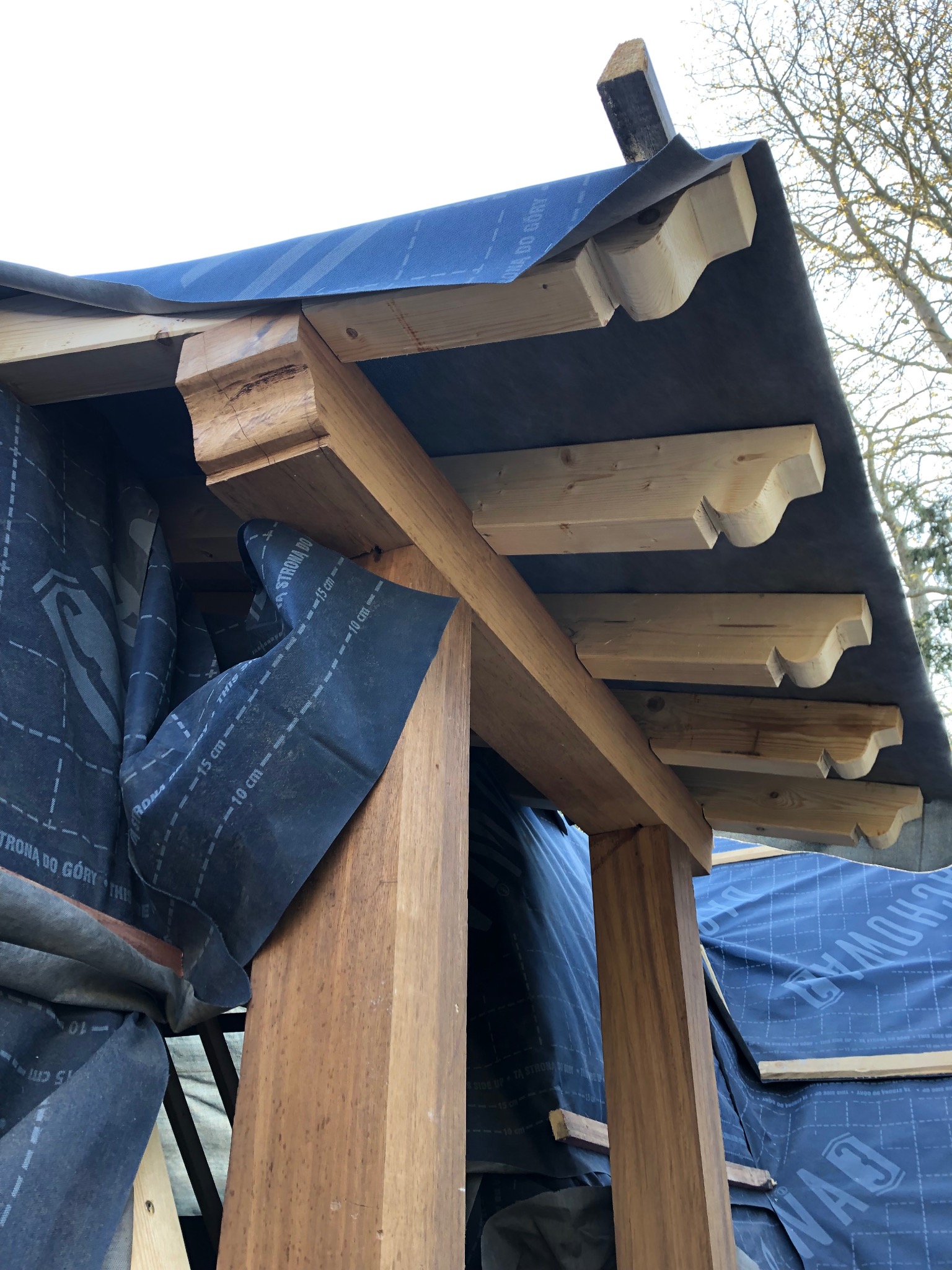 Konstrukcja dachu w trakcie budowy, widoczne drewniane elementy nośne i membrana dachowa z nadrukiem, jasne drewno krokwi dekoracyjnych.
