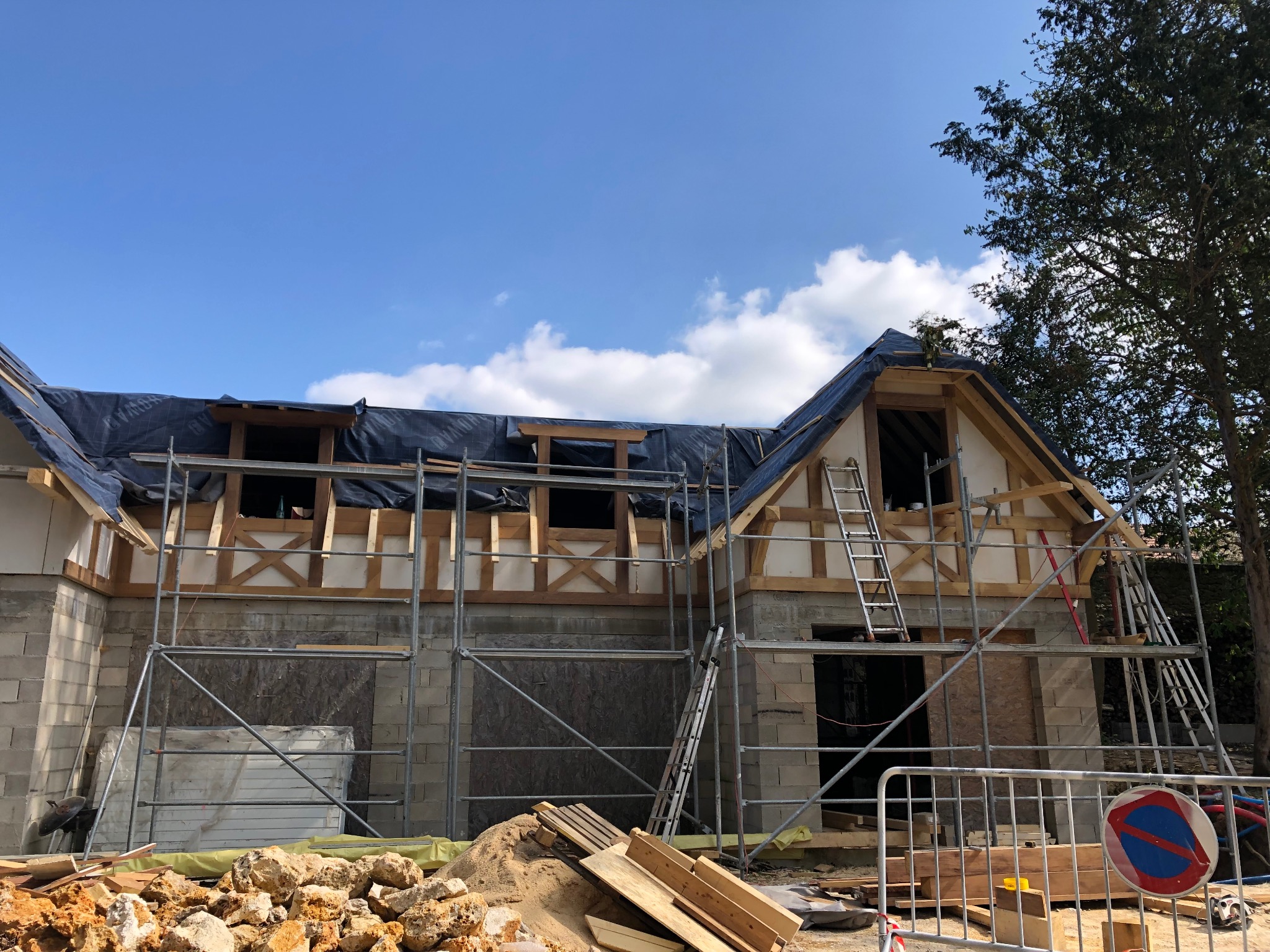 Wznoszenie konstrukcji domu z elementami drewnianymi, widoczne rusztowania, materiały budowlane i częściowo ukończona ściana z bloczków.