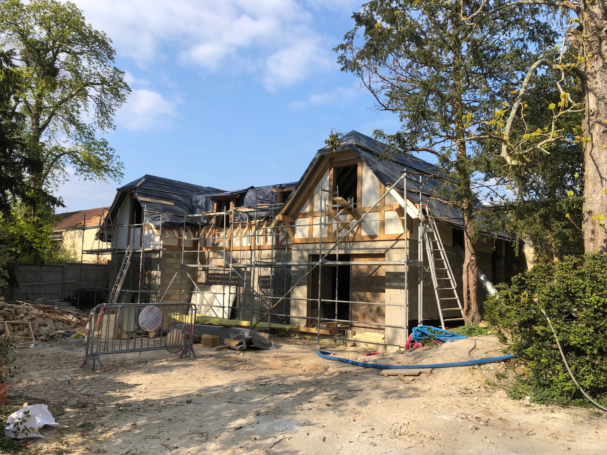 Wznoszenie dwukondygnacyjnego domu z drewnianą konstrukcją szkieletową, widoczne rusztowania, zabezpieczenie dachu folią, teren budowy w trakcie prac.