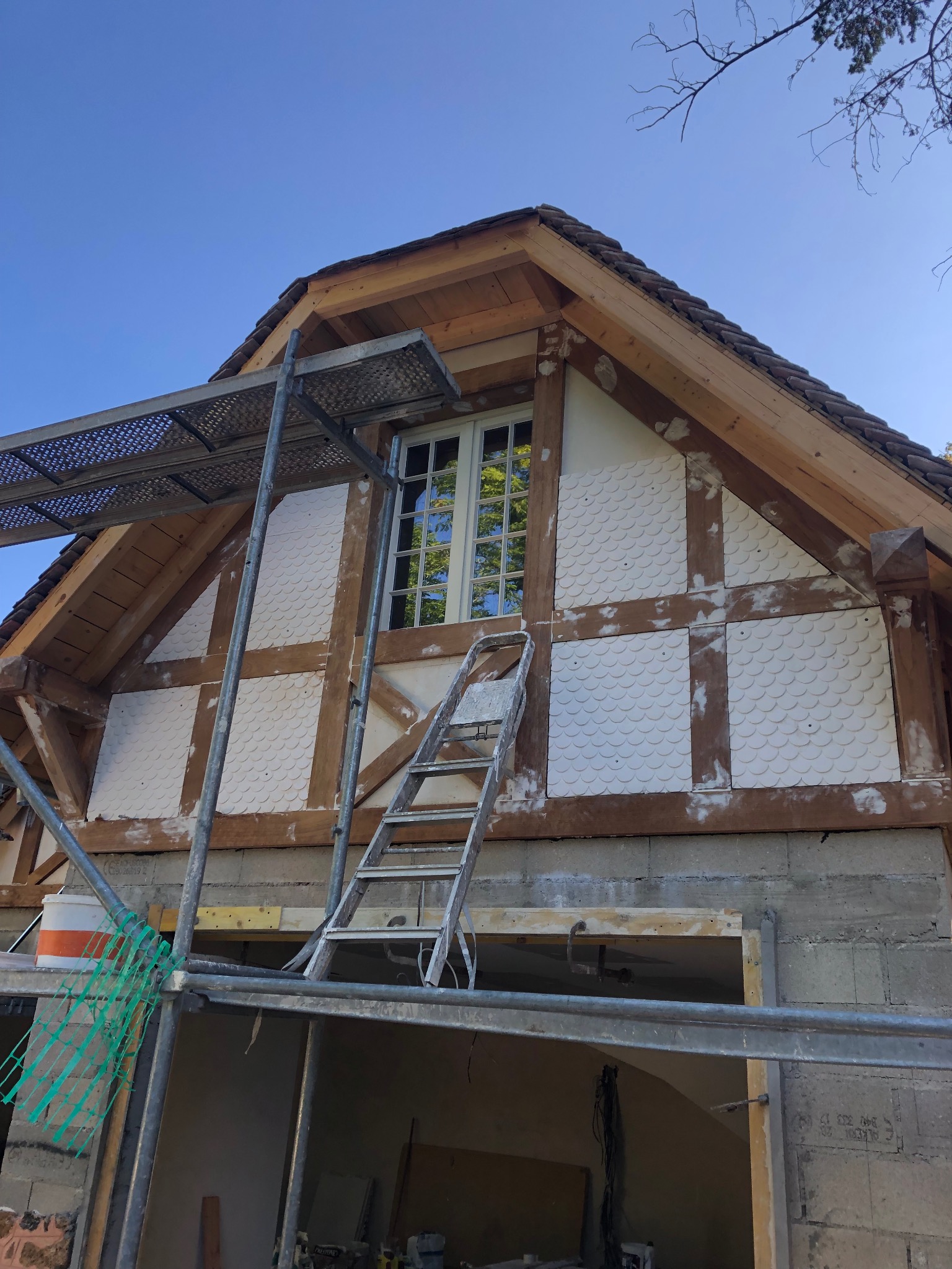 Wznoszona konstrukcja budynku z drewnianym szkieletem, częściowo otynkowana, z widocznym rusztowaniem i drabiną, na tle błękitnego nieba.