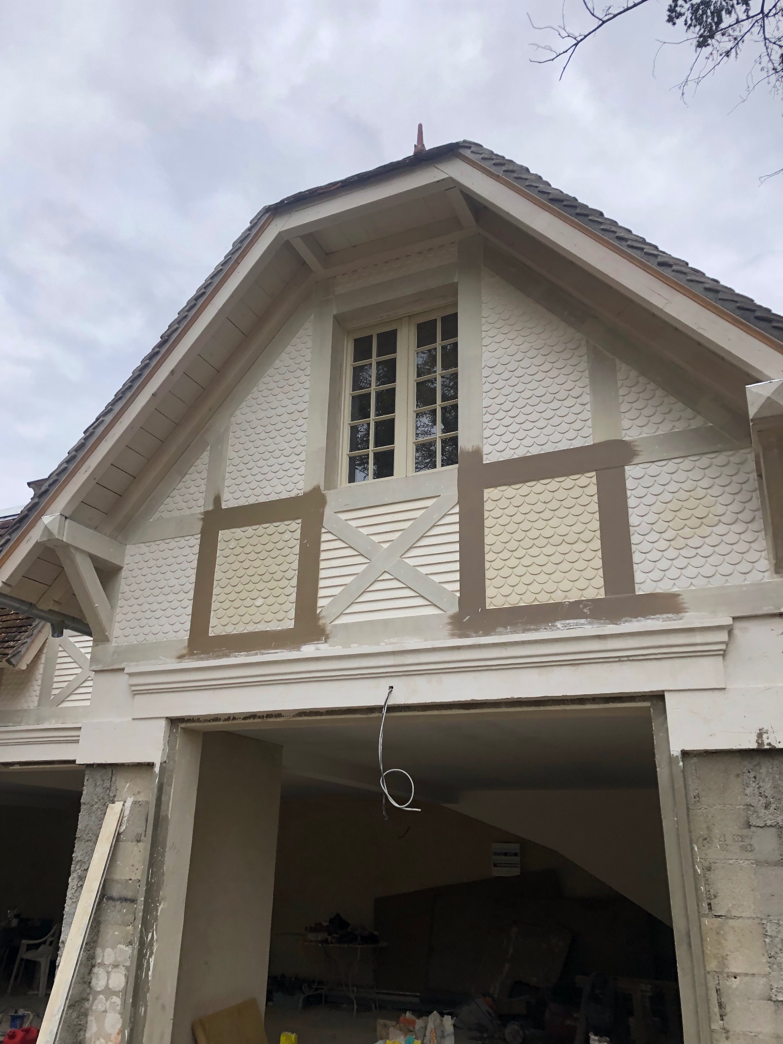 Fragment elewacji budynku z charakterystyczną dekoracją w formie łusek, widoczne elementy konstrukcyjne i okno, prace wykończeniowe w toku.