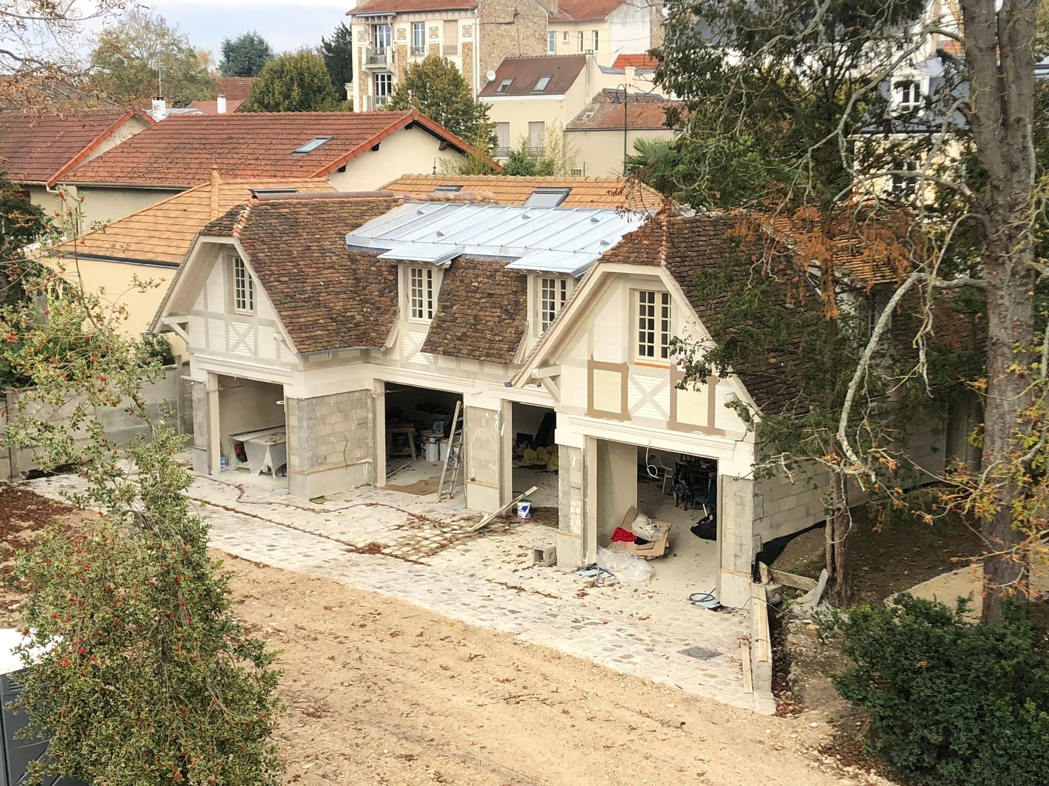 Wznoszenie konstrukcji garażu z elementami muru pruskiego, widoczne surowe ściany, dach z dachówki i blachy, teren budowy z kostką brukową i ziemią.