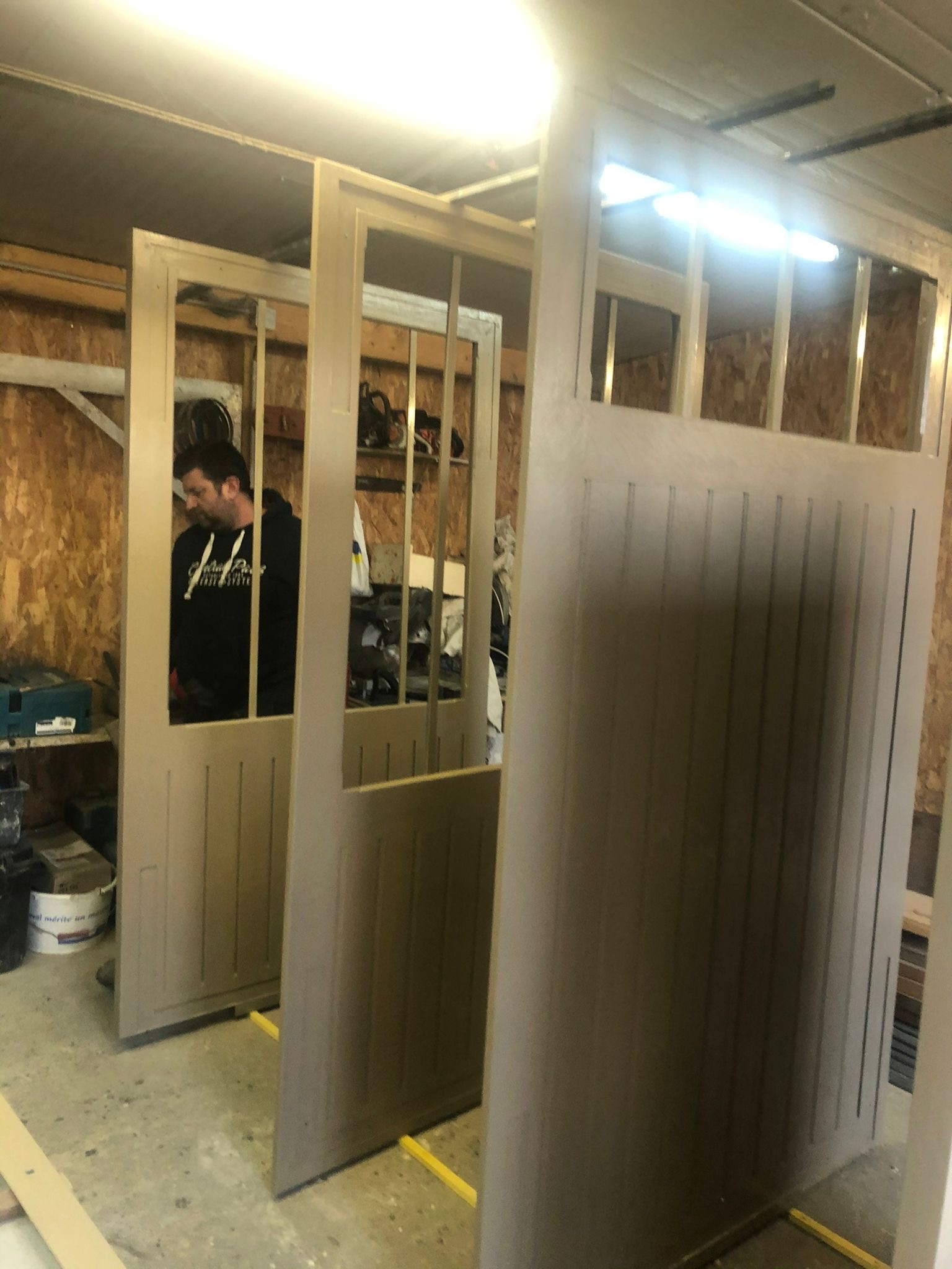 Wnętrze warsztatu z pracownikiem montującym elementy szkieletowe ścian z oknami, widoczne narzędzia i materiały budowlane.