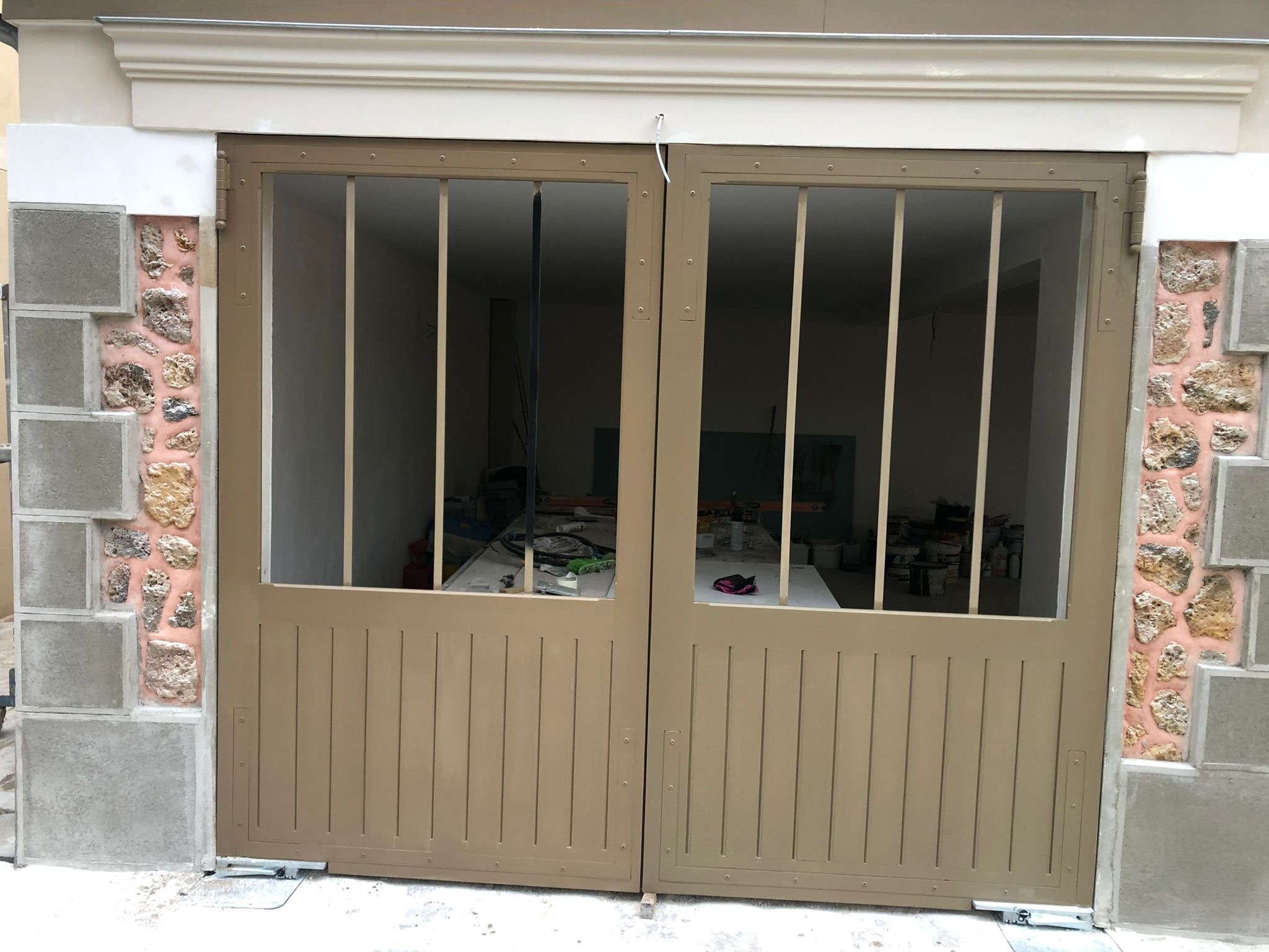 Dwuskrzydłowa, metalowa brama w kolorze beżowym, z pionowymi prętami w górnej części, zamontowana w kamiennym murze z elementami betonowymi, widok na wnętrze budynku w trakcie prac wykończeniowych.