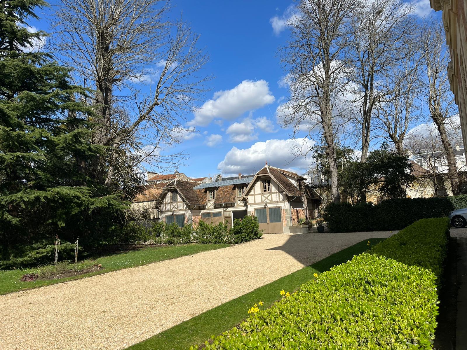 Widok z zewnątrz na budynek z elementami szkieletowymi i dwuspadowym dachem, otoczony zielenią i żwirową ścieżką w słoneczny dzień.