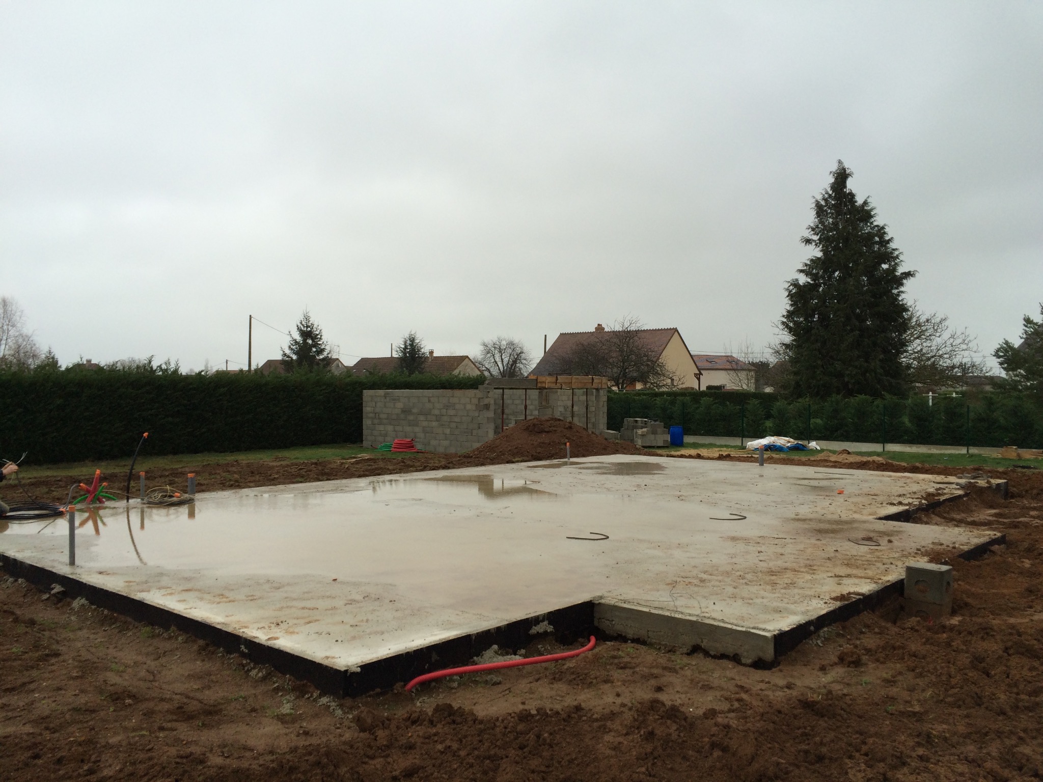 Wylany, lekko wilgotny fundament płyty betonowej z wystającymi rurami instalacyjnymi, obok widoczny fragment ściany fundamentowej z bloczków betonowych, w tle zabudowa jednorodzinna i pochmurne niebo.