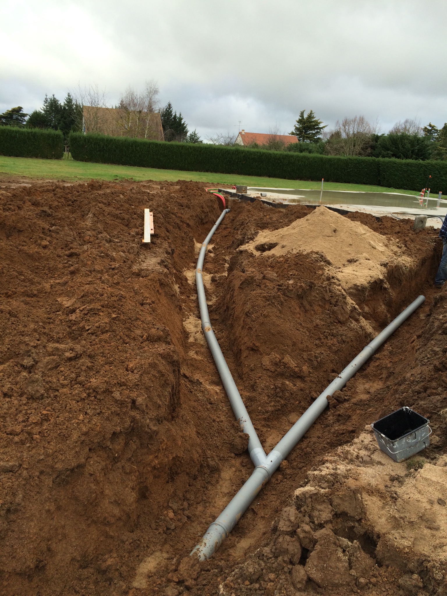 Wykop pod instalacje kanalizacyjne z rurami PVC w kształcie litery Y, poziomica na krawędzi wykopu, w tle trawnik i domy.