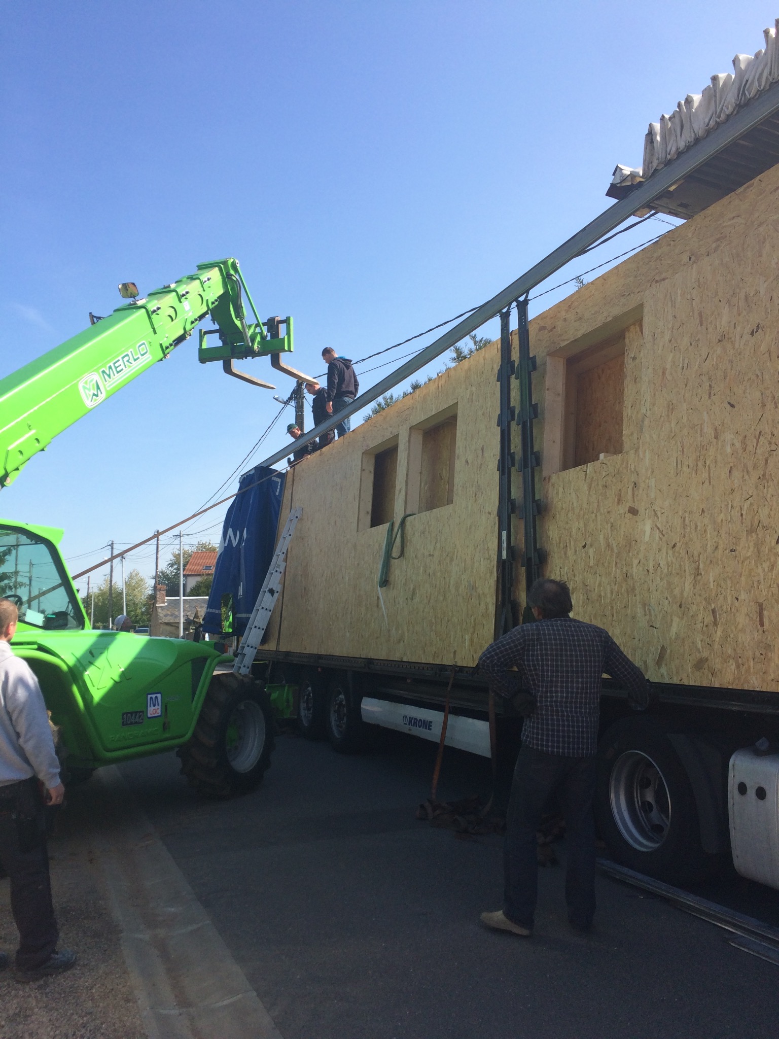Transport modułu domu z wyciętymi oknami na naczepie ciężarówki, podnoszony przez zielony teleskopowy wózek widłowy Merlo, robotnicy na module i obok naczepy.