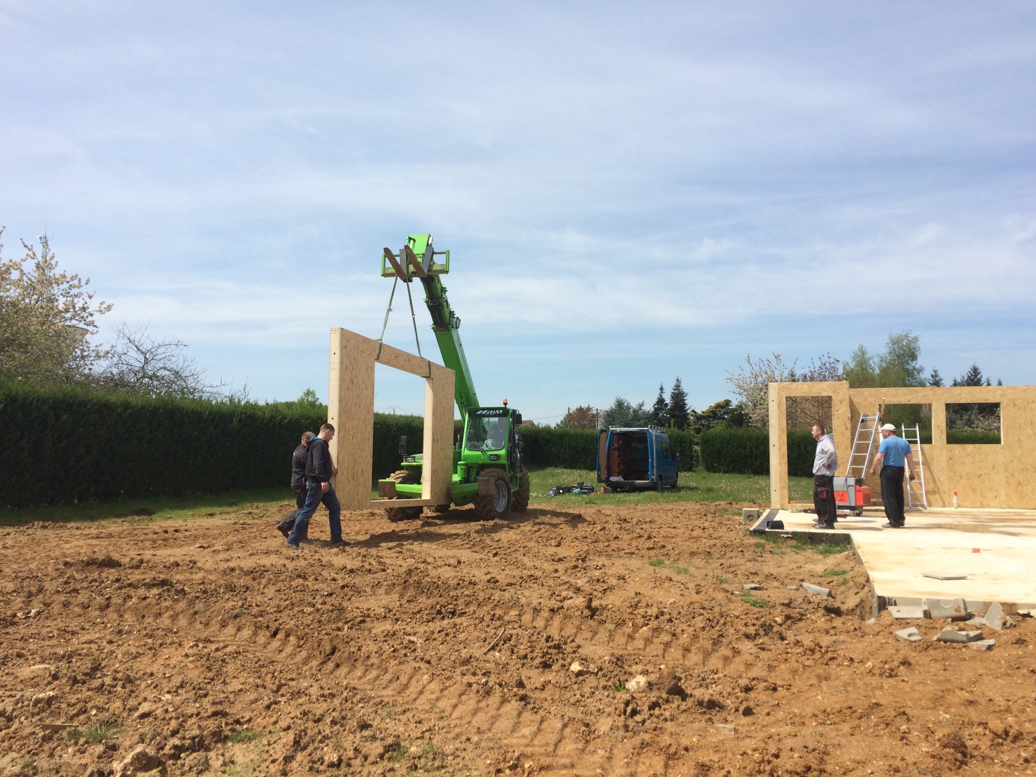 Montaż ściany z drewna na fundamencie domu, z użyciem podnośnika teleskopowego; dwóch mężczyzn przenosi element, w tle widoczny bus i zielony żywopłot.
