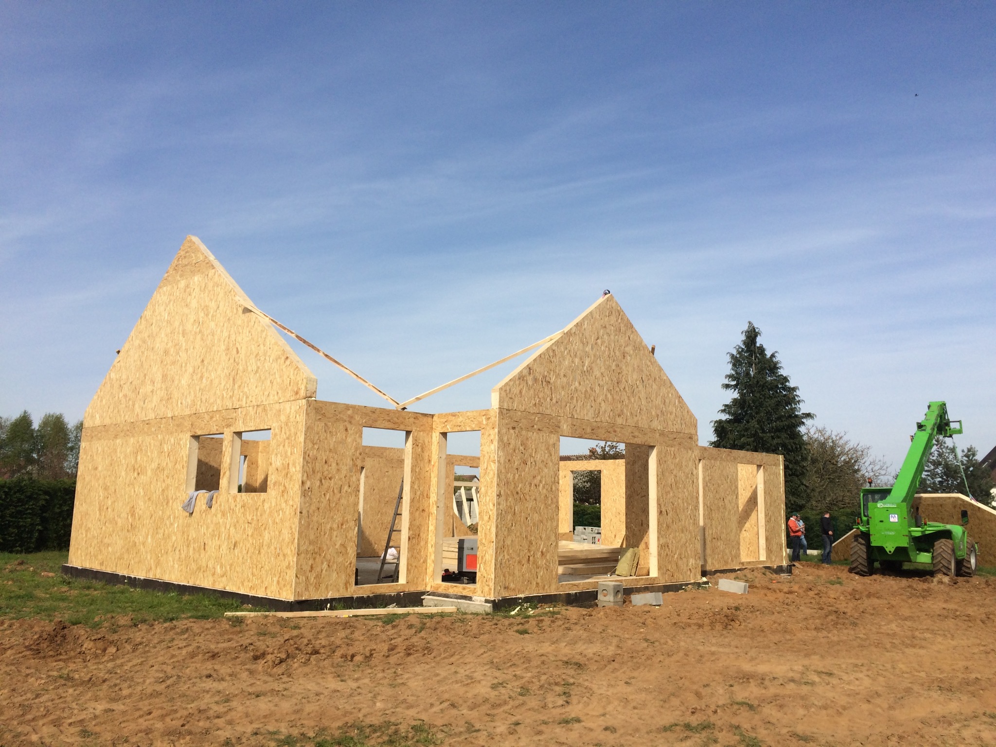 Wznoszenie konstrukcji domu z płyt drewnopochodnych, widoczny podnośnik teleskopowy i pracownicy w tle.