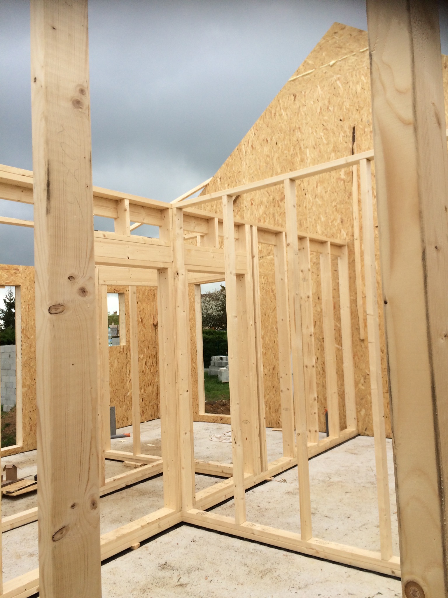 Wnętrze domu w trakcie budowy, widoczne drewniane elementy konstrukcyjne ścian i dachu z płyt OSB, betonowa podłoga, w tle pochmurne niebo.