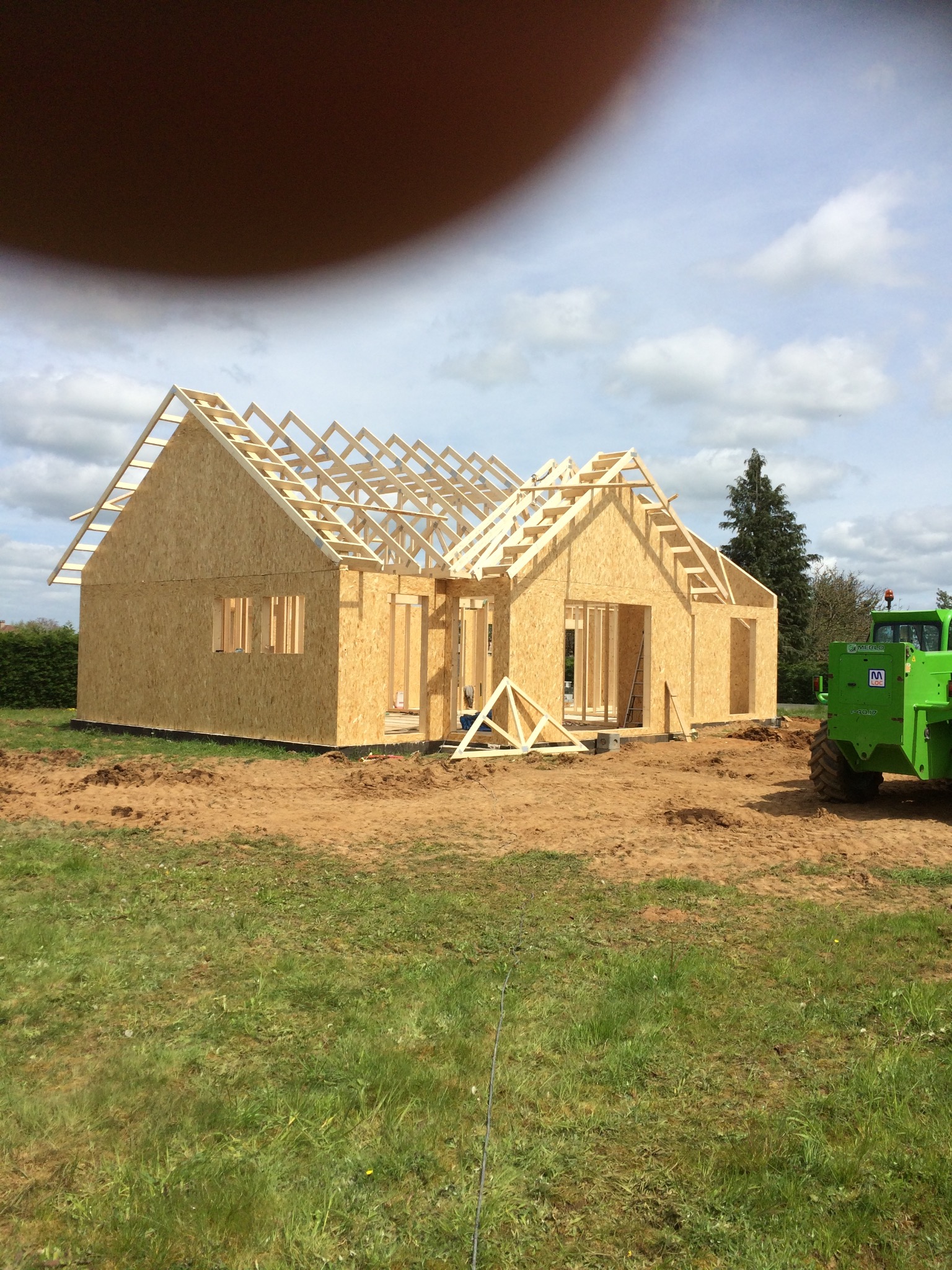 Wznoszenie konstrukcji domu z drewnianych paneli OSB, widoczne elementy więźby dachowej i zielony podnośnik teleskopowy Merlo w tle.