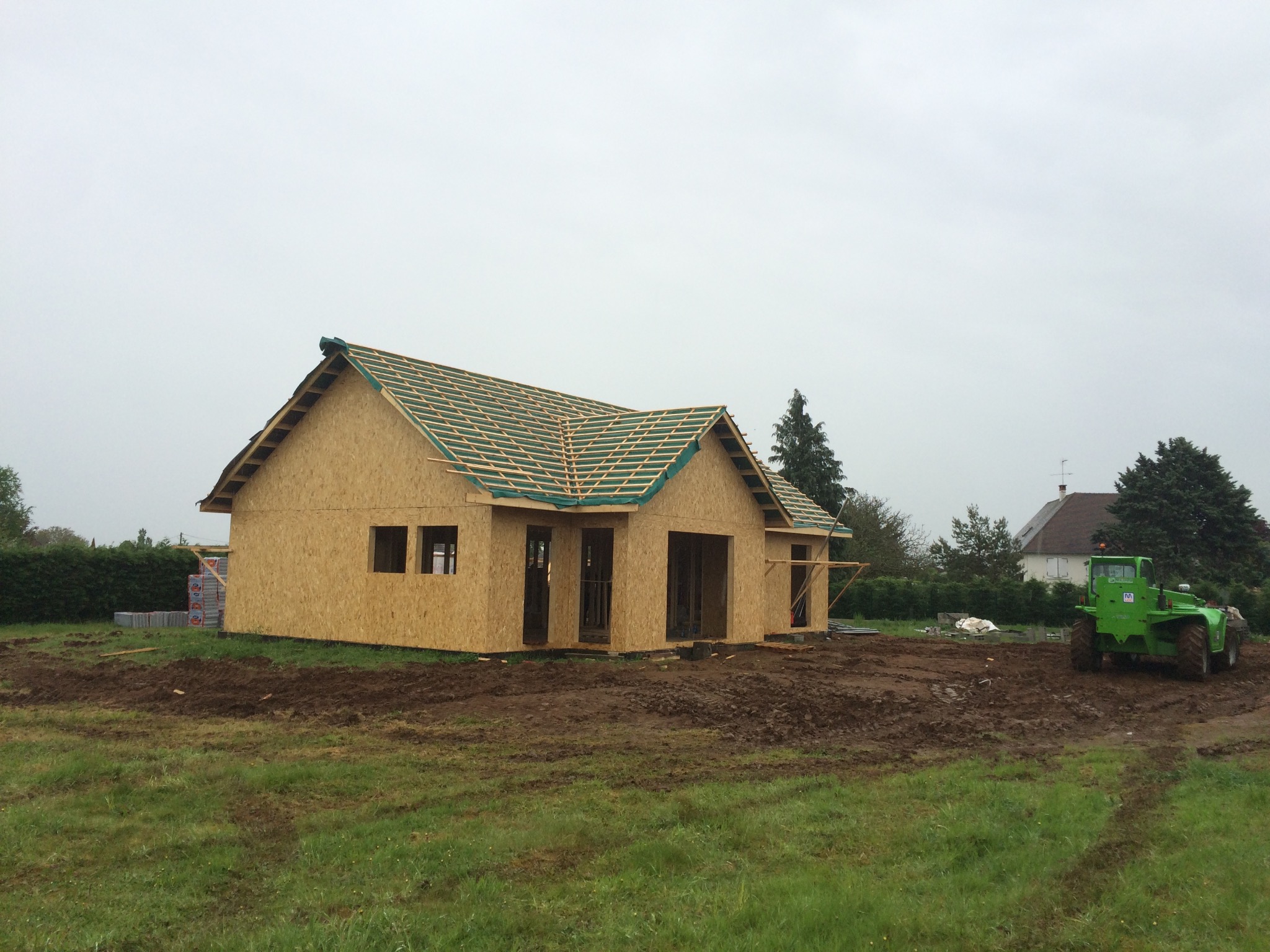 Budowa domu szkieletowego: surowa konstrukcja z płyty OSB, widoczne krokwie dachowe z zieloną folią, traktor na błotnistym terenie w tle.
