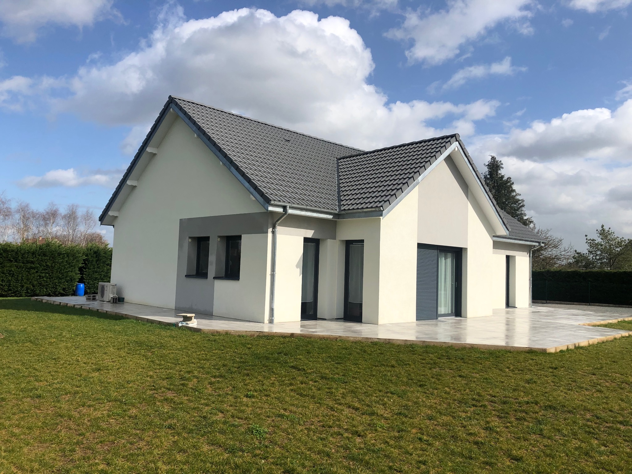 Nowoczesny, parterowy dom z szarą dachówką i jasną elewacją, otoczony trawnikiem i częściowo wyłożony płytami.