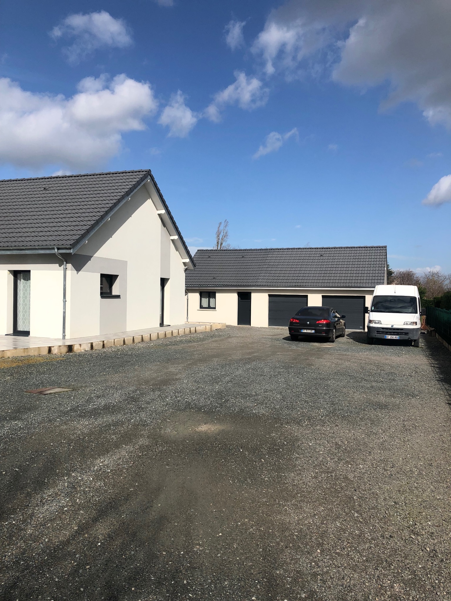 Nowoczesny dom jednorodzinny z szarym dachem i jasną elewacją, widok z zewnątrz na podjazd z szarego żwiru, zaparkowane samochody osobowe i dostawcze, błękitne niebo z chmurami.