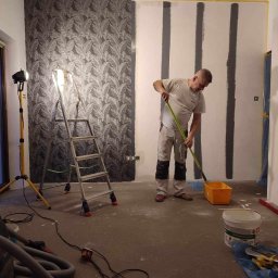 Pogotowie Remontowe Radosław Świderski - Mężczyzna maluje pionowe pasy na ścianie pokoju za pomocą wałka, obok stoi drabina i lampa halogenowa, na podłodze widoczne akcesoria malarskie i odkurzacz.