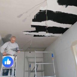 Pogotowie Remontowe Radosław Świderski - Mężczyzna na drabinie szpachluje sufit w pomieszczeniu, widoczne niedokończone pasy czarnej farby i wisząca żarówka.