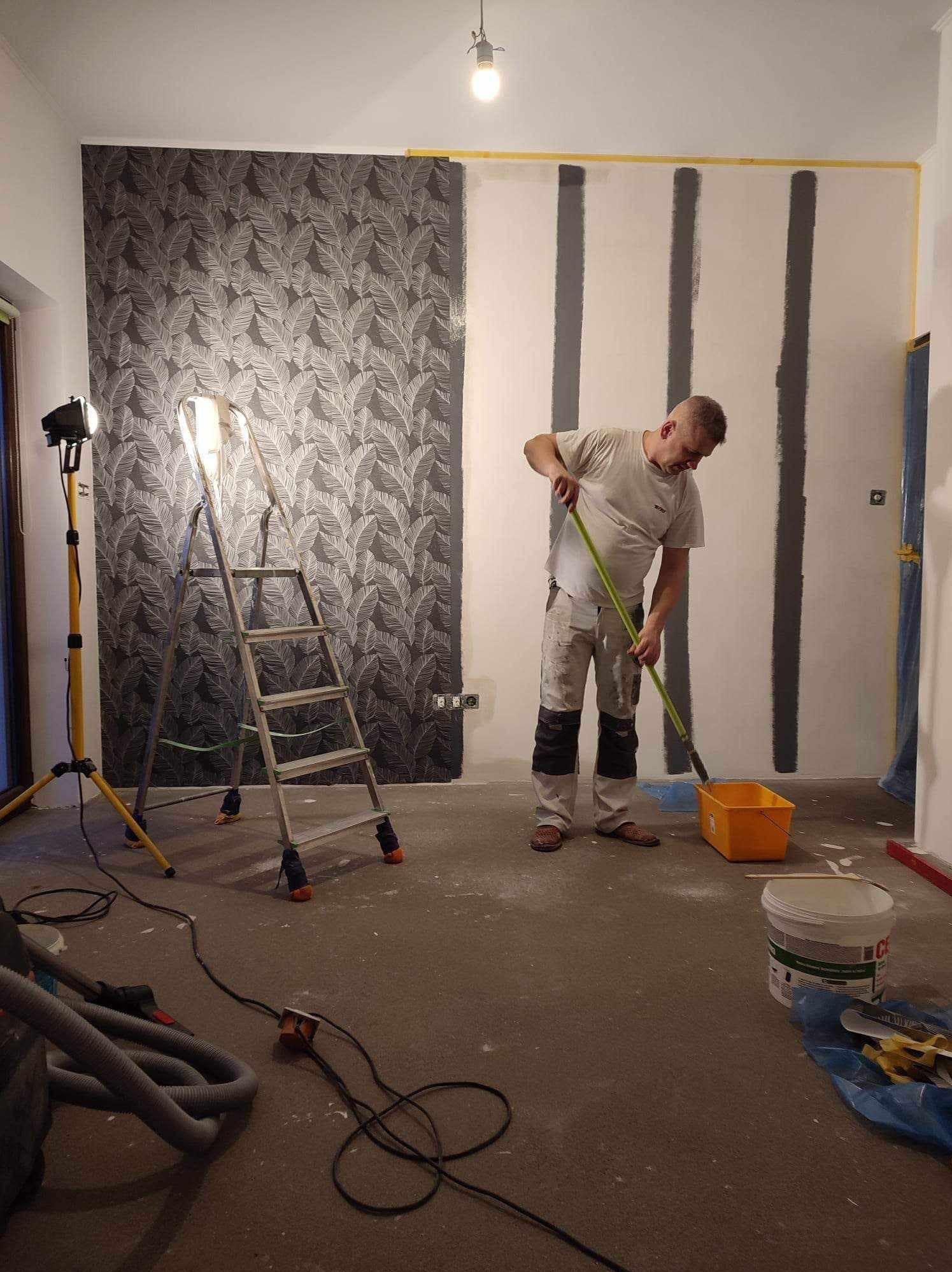 Mężczyzna maluje pionowe pasy na ścianie pokoju za pomocą wałka, obok stoi drabina i lampa halogenowa, na podłodze widoczne akcesoria malarskie i odkurzacz.