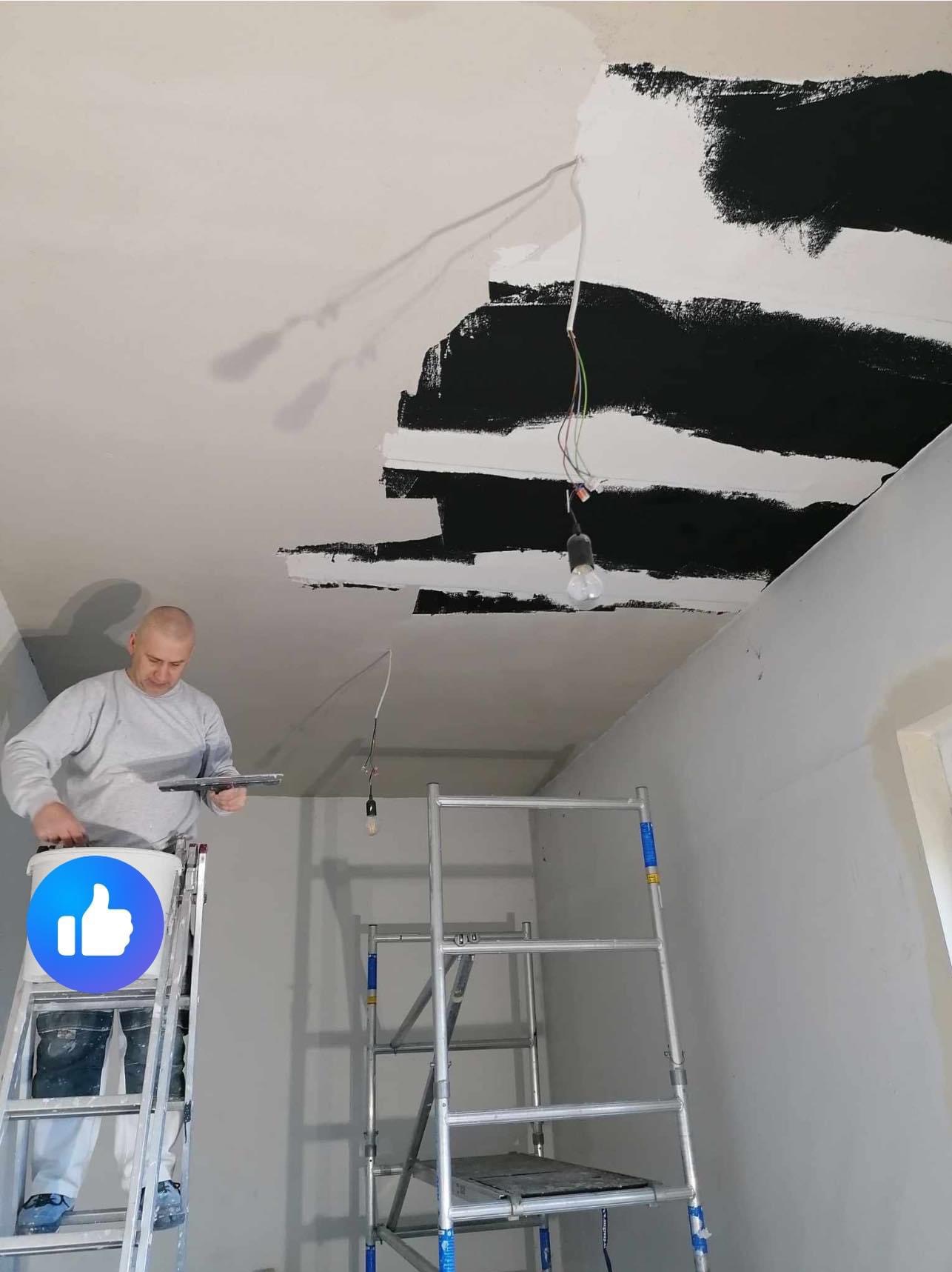 Mężczyzna na drabinie szpachluje sufit w pomieszczeniu, widoczne niedokończone pasy czarnej farby i wisząca żarówka.