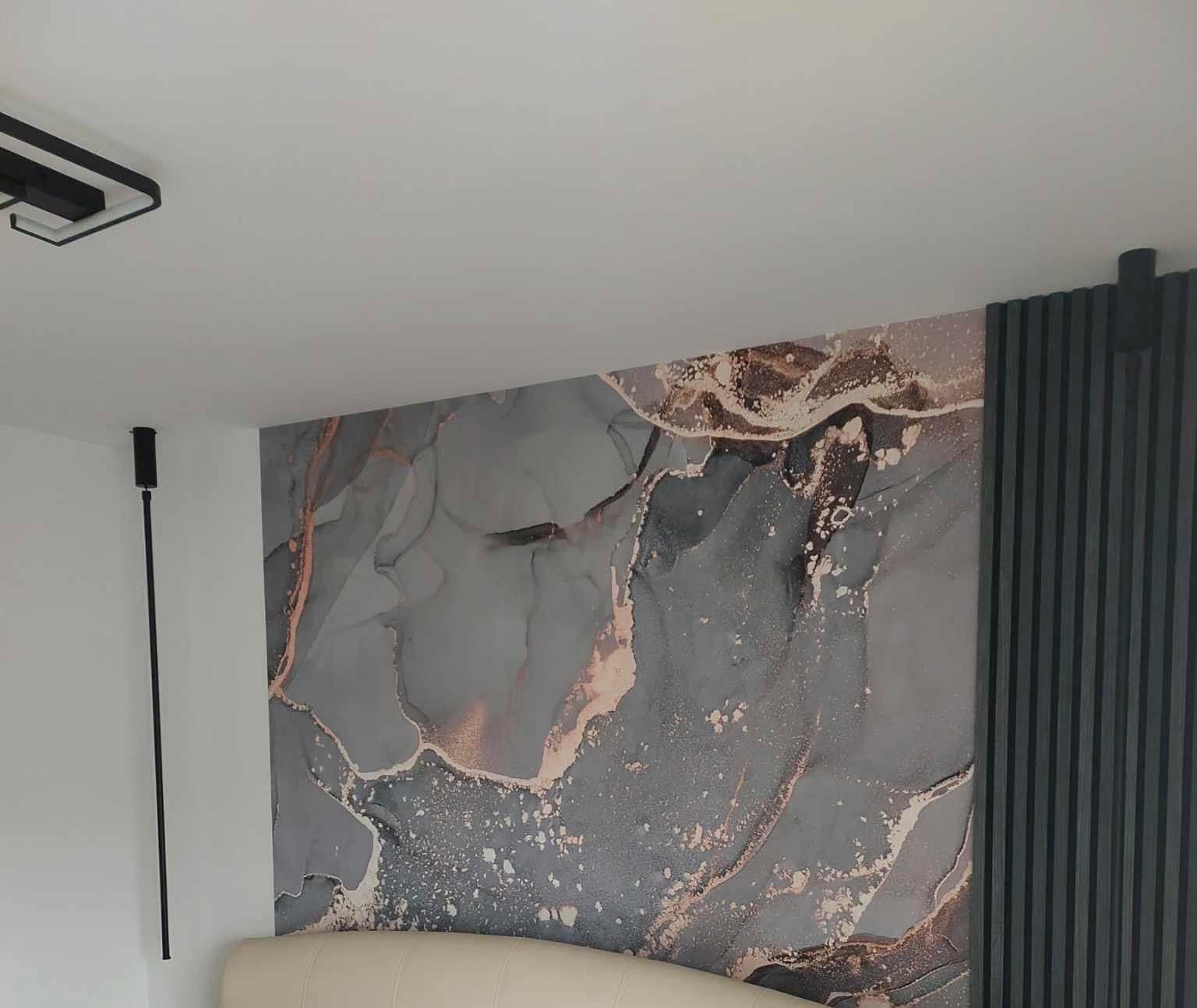 Ściana w sypialni z abstrakcyjną tapetą w odcieniach szarości i złota, sąsiadująca z ciemnoszarymi panelami dekoracyjnymi, uzupełniona o minimalistyczne oświetlenie.