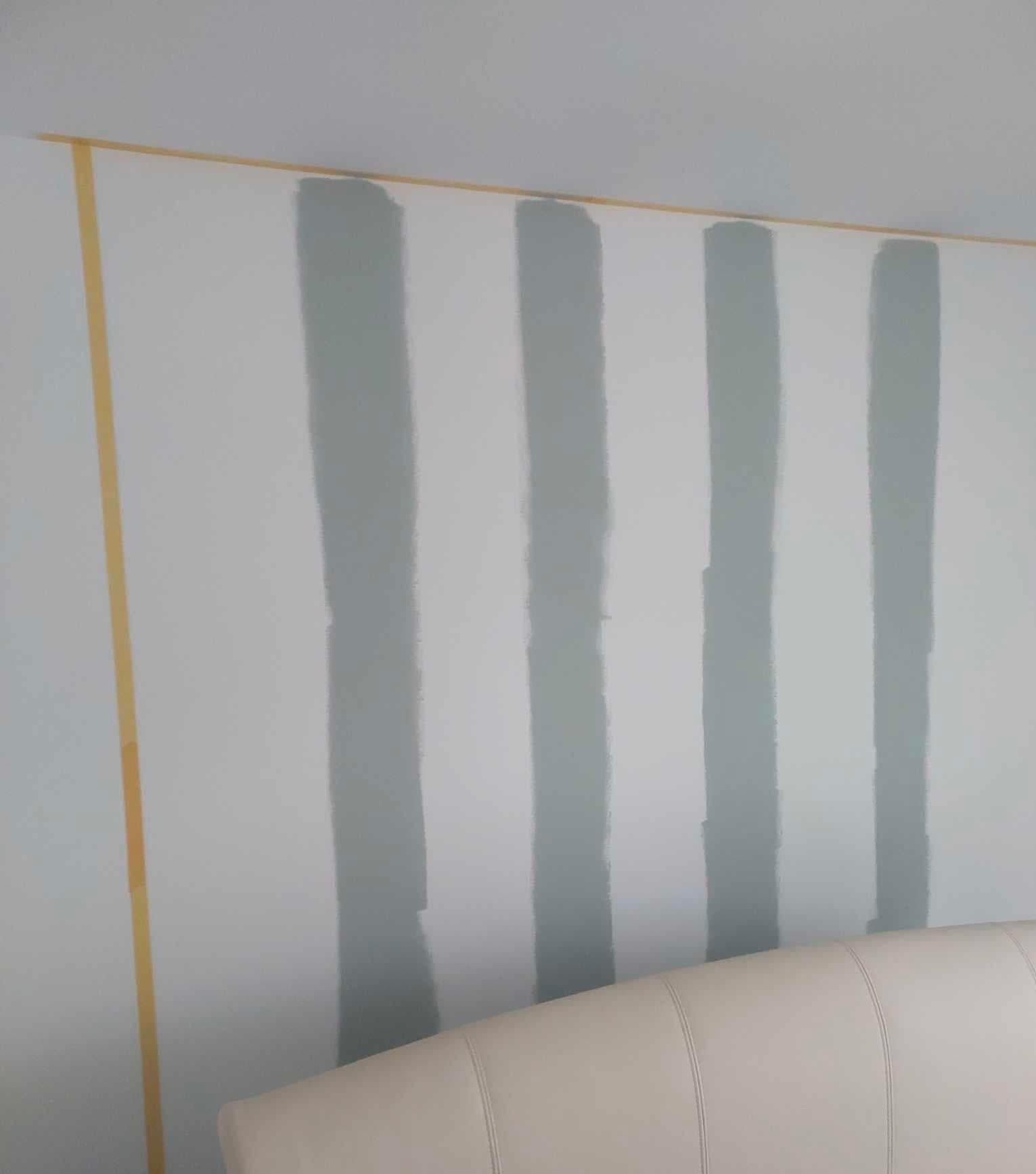 Ściana z pionowymi pasami w odcieniach szarości, oddzielona taśmą malarską, widoczny fragment wezgłowia łóżka.