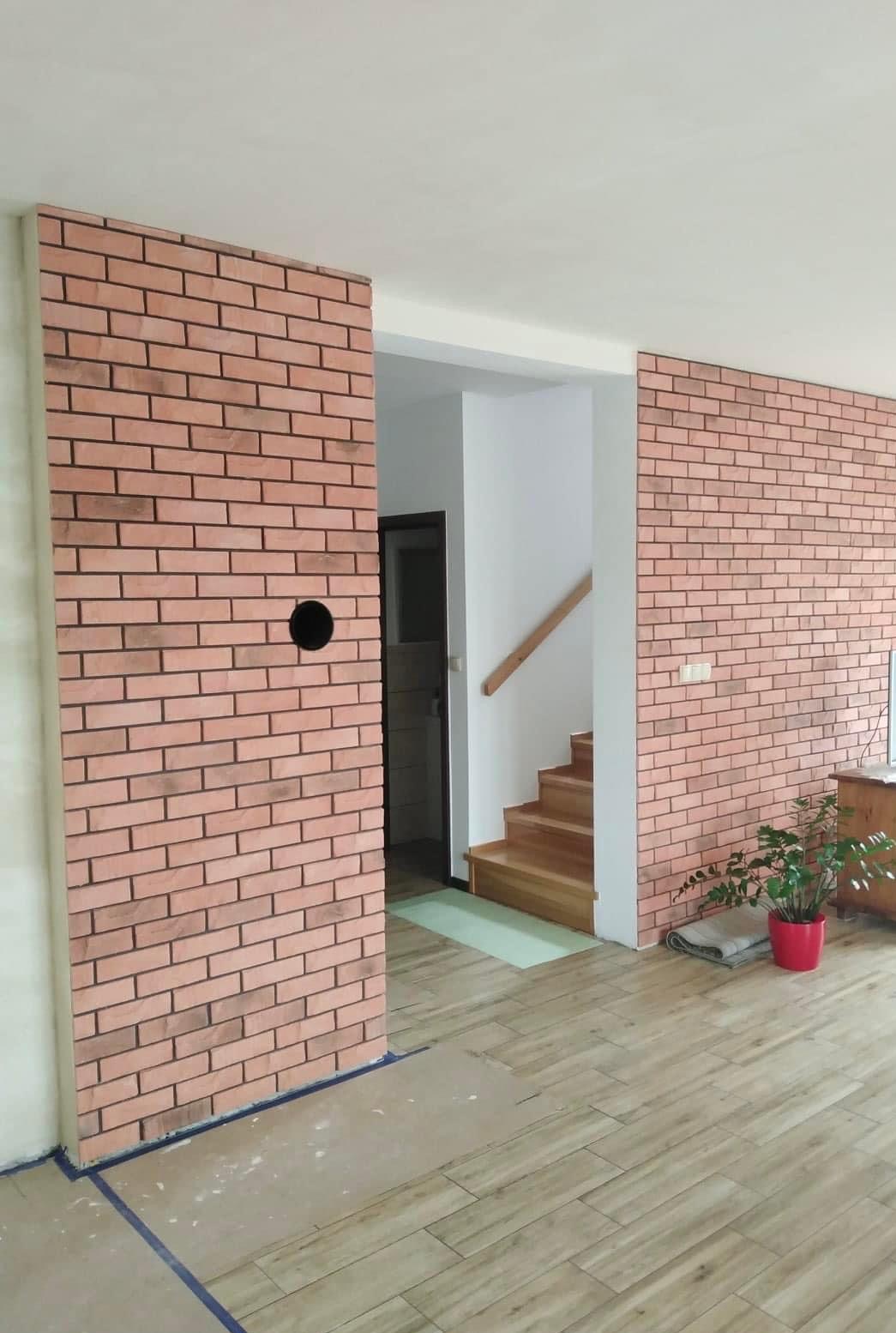 Wykończona ściana z cegły dekoracyjnej w salonie, widoczny otwór na gniazdko elektryczne, drewniane schody w tle i podłoga wyłożona panelami imitującymi drewno.